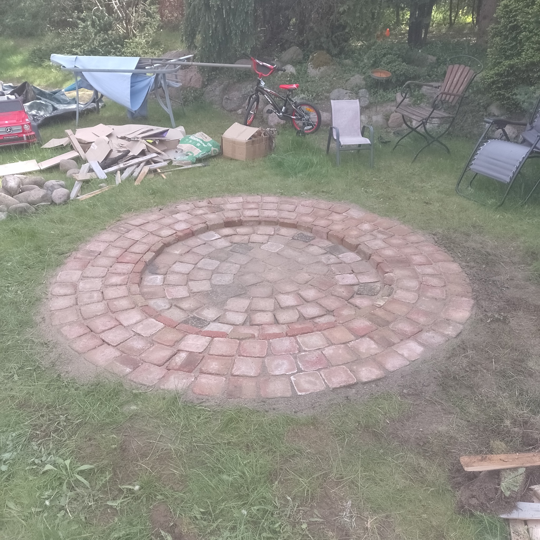 Okrągły plac z czerwonej kostki w trawie, przygotowany pod ognisko, w tle porozrzucane narzędzia i materiały budowlane, rower i meble ogrodowe.