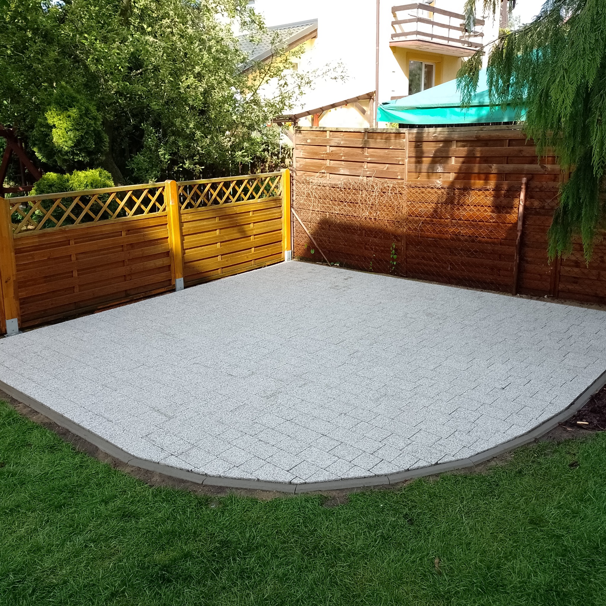 Wykończony plac z jasnoszarej kostki brukowej ułożonej w regularny wzór, obrzeża z szarego betonu, ogrodzenie drewniane w tle, trawnik wokół.