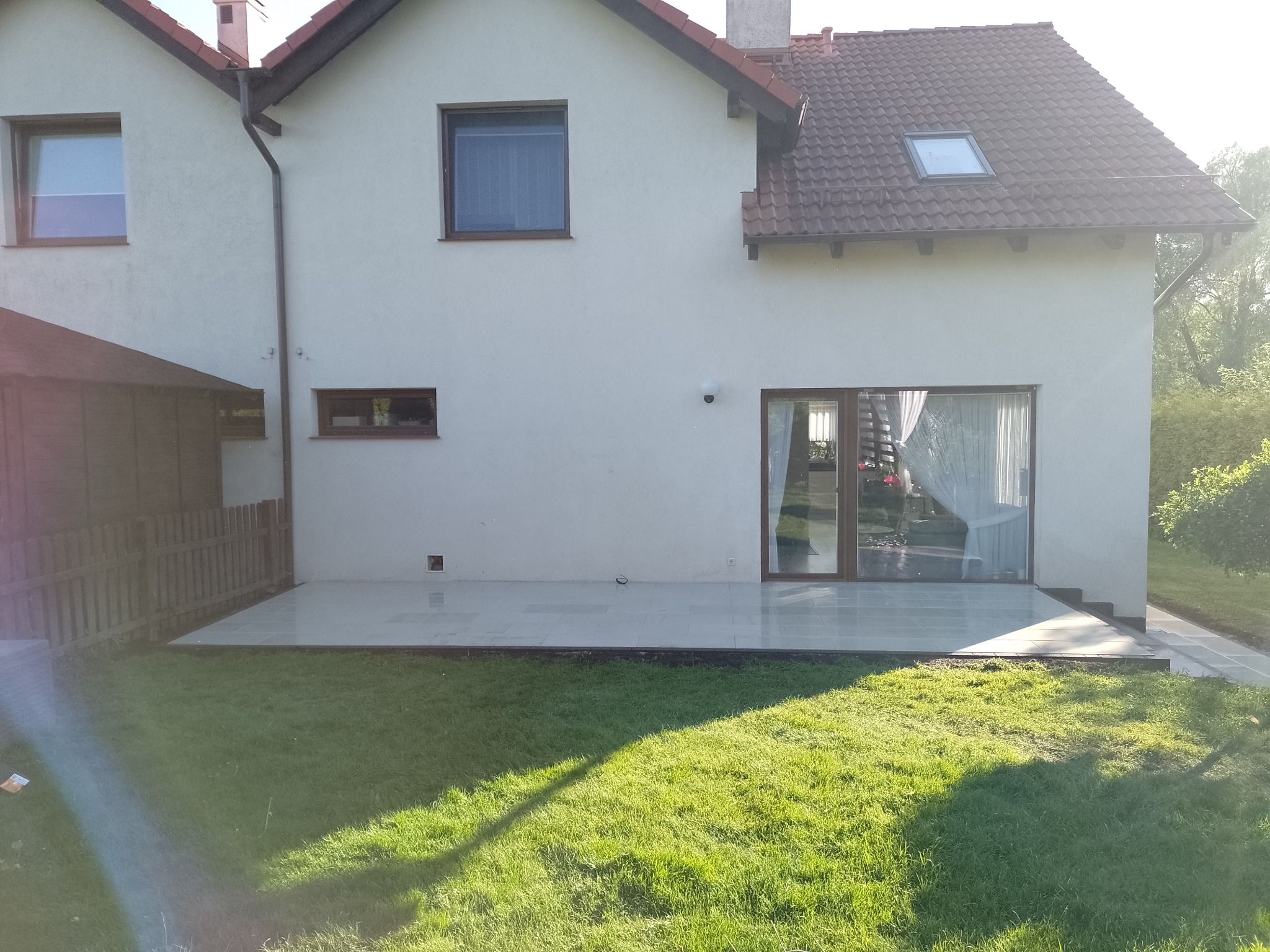 Jasne płytki tarasowe przylegające do domu z przesuwnymi drzwiami, widok na trawnik i częściowo ogrodzenie z drewnianych sztachet.