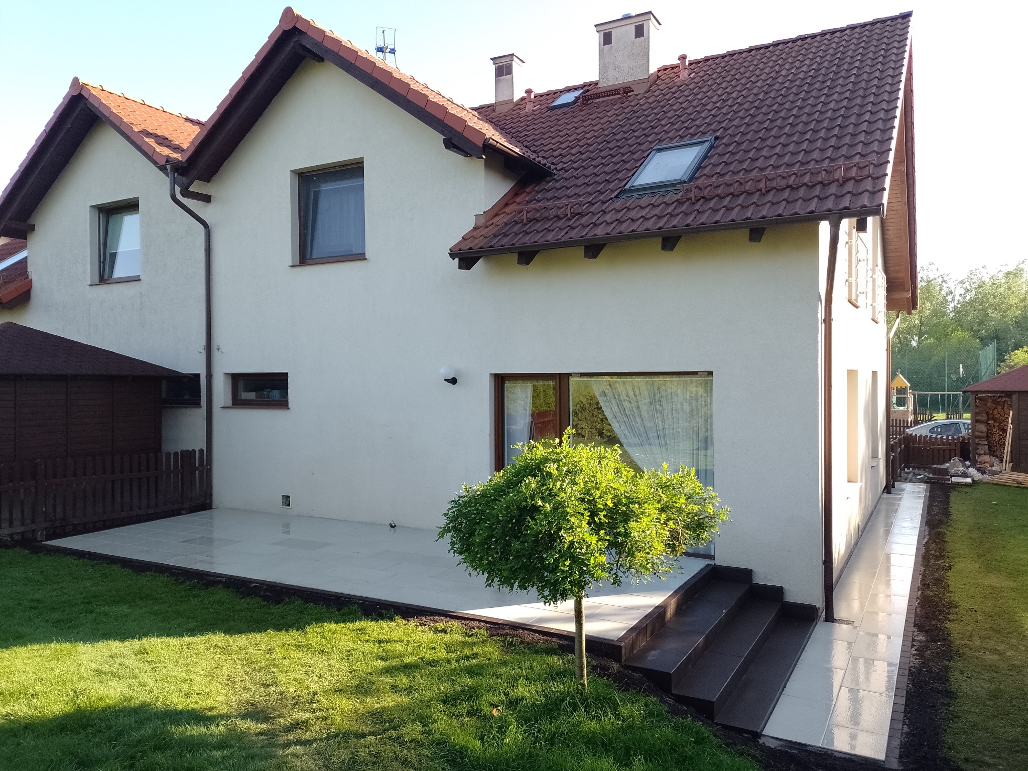 Jasnoszara, ułożona kostka tworzy taras i ścieżkę wokół domu z beżową elewacją i czerwoną dachówką, widoczna trawa i małe drzewko przed oknem.