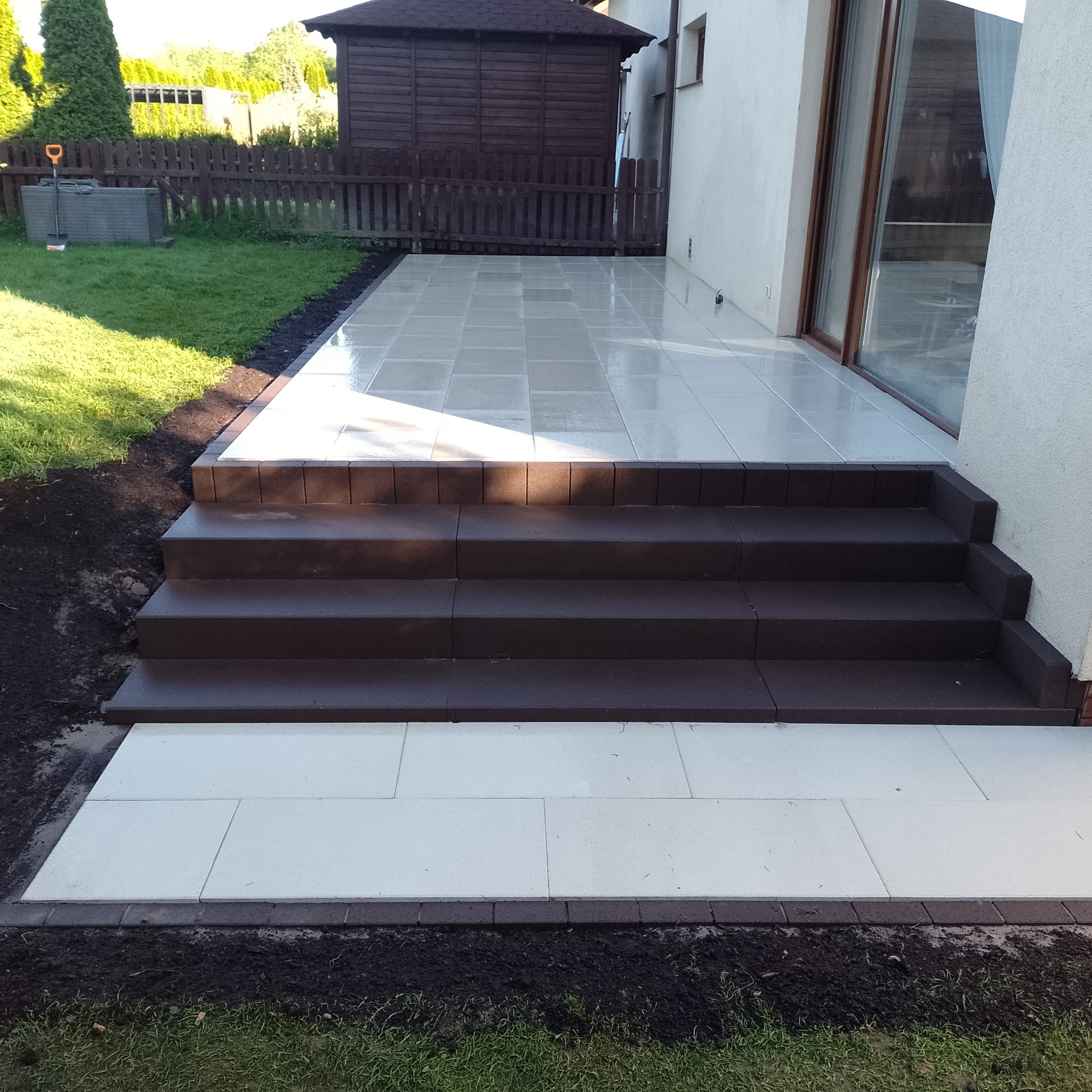 Nowo wyłożony taras z jasnych, kwadratowych płyt, prowadzący do domu po trzech ciemnobrązowych stopniach, obrzeża z ciemnej kostki, świeżo obsypana ziemia przy trawniku.