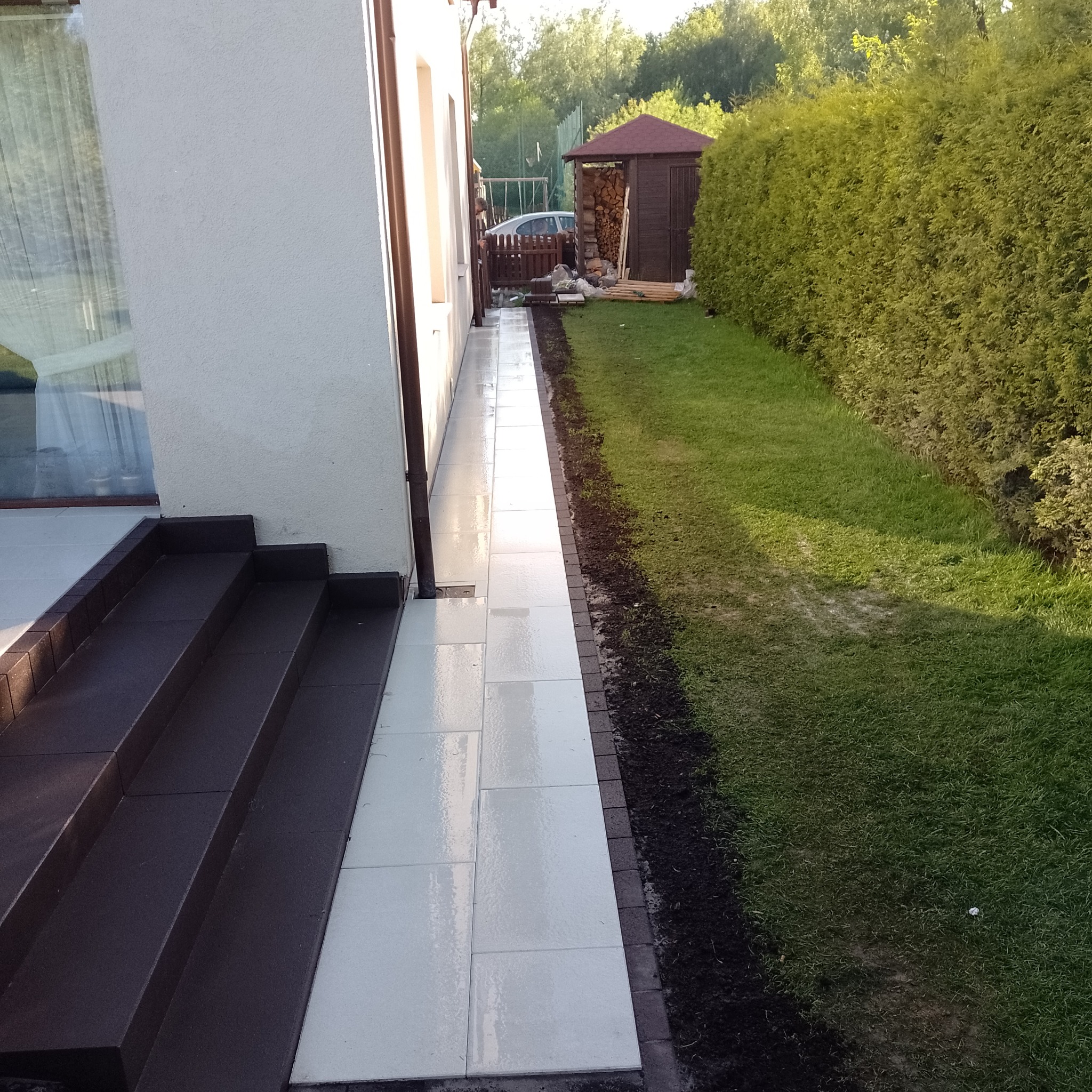 Ułożona, jasnoszara kostka brukowa tworząca ścieżkę wzdłuż ściany domu, z ciemnymi schodami wejściowymi i trawnikiem z żywopłotem w tle.
