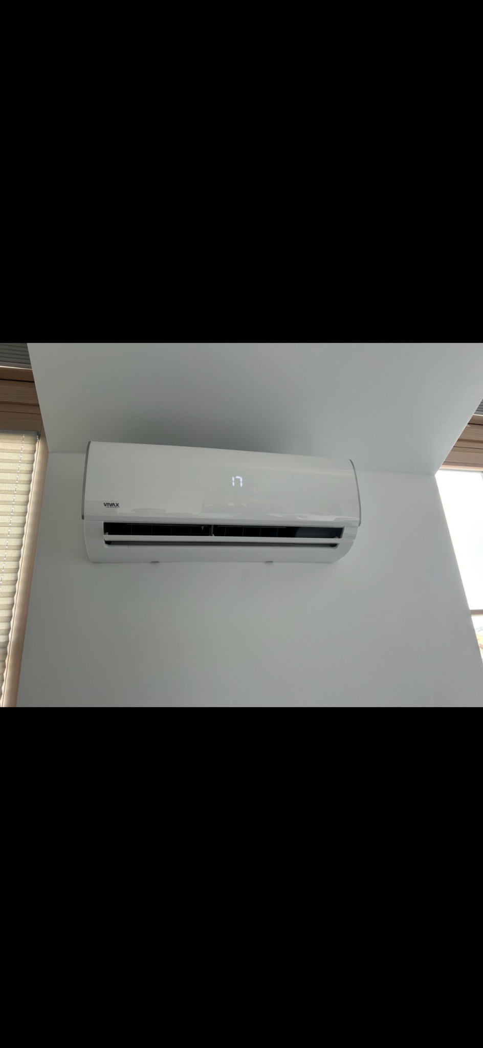 Biały klimatyzator marki Vivax zamontowany na białej ścianie w pomieszczeniu z żaluzjami w tle, wyświetlający temperaturę 17 stopni.
