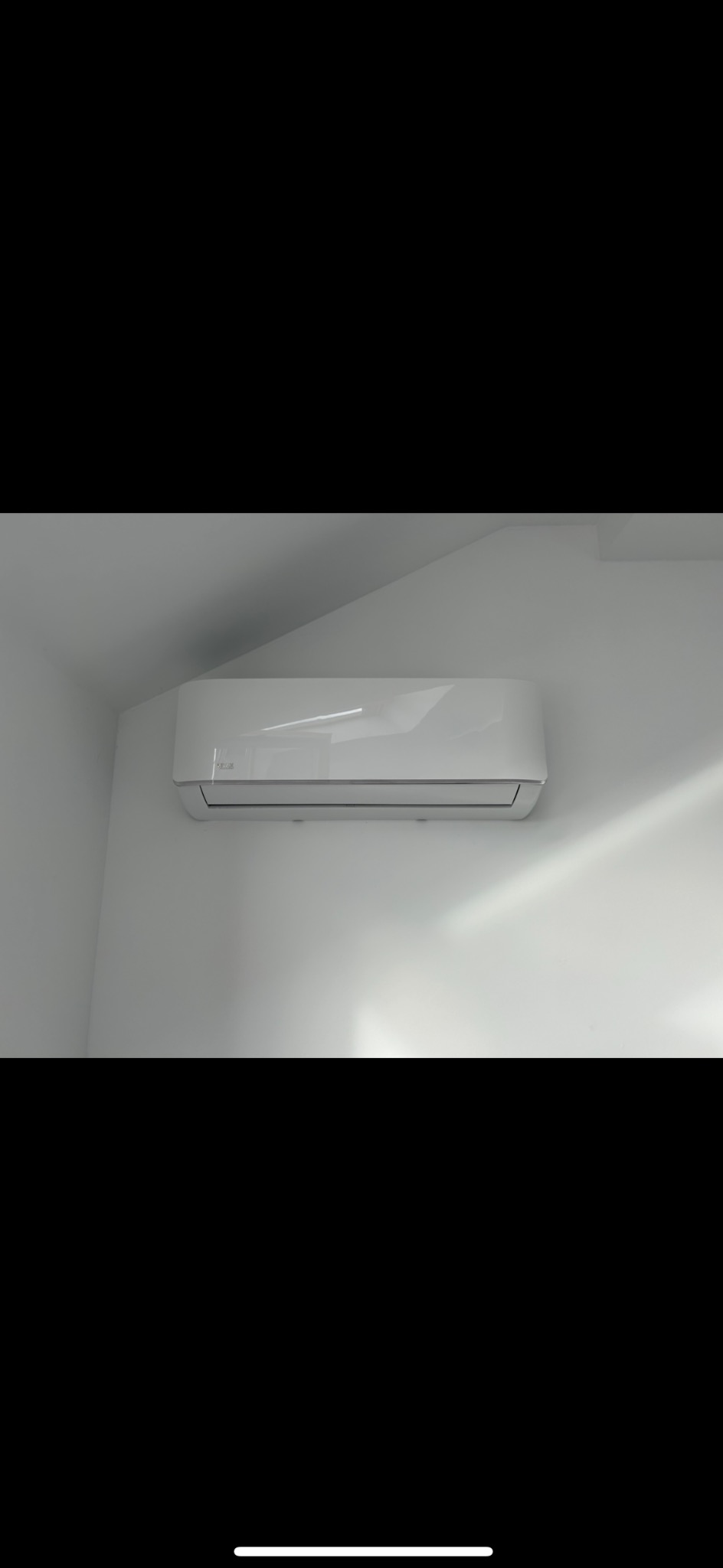 Biała jednostka klimatyzacyjna zamontowana na białej ścianie w rogu pomieszczenia, widok od dołu, delikatne światło.