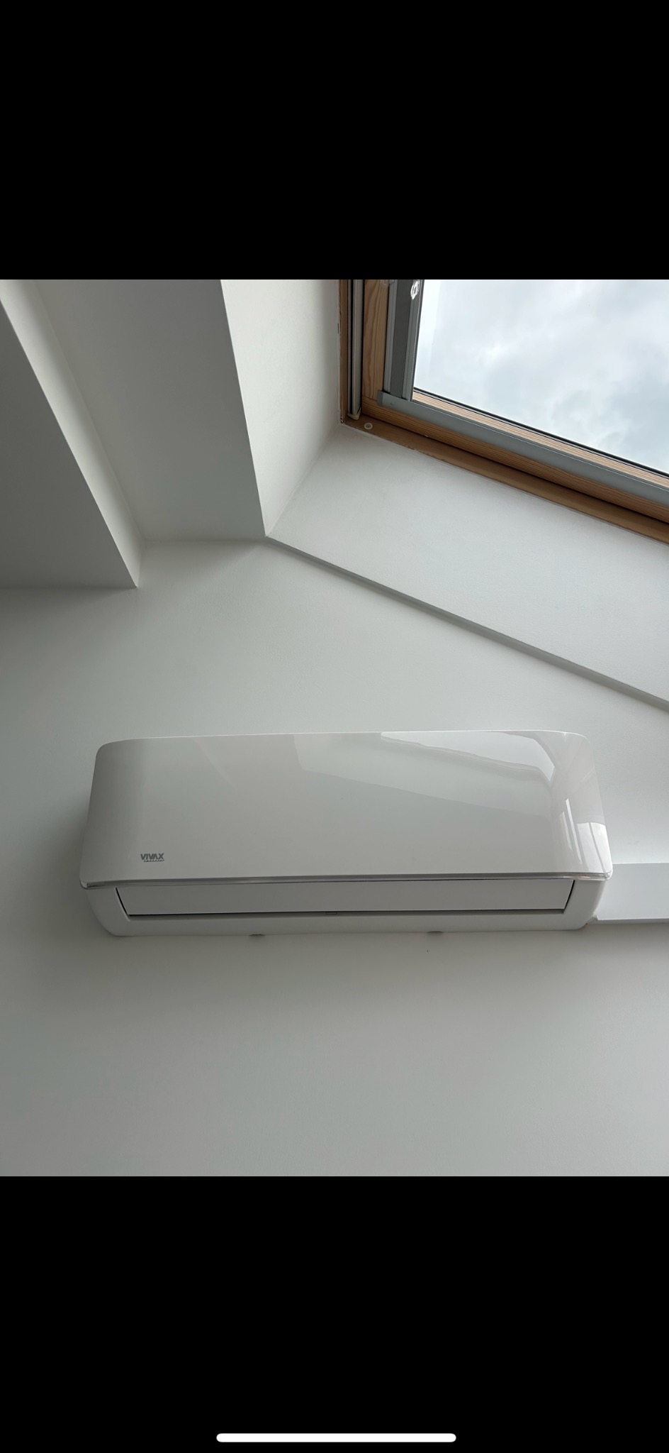 Biała klimatyzacja VIVAX zamontowana na białej ścianie pod oknem dachowym w jasnym pomieszczeniu.