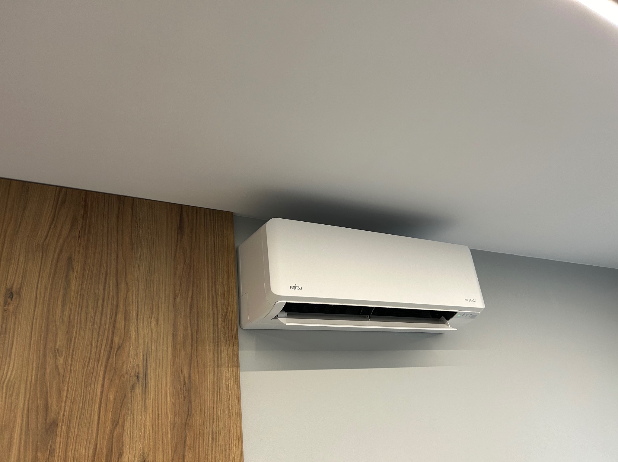 Biała jednostka klimatyzacji Fujitsu Airstage zamontowana na szarej ścianie, widoczna z boku, z fragmentem drewnianej okładziny ściany po lewej stronie.