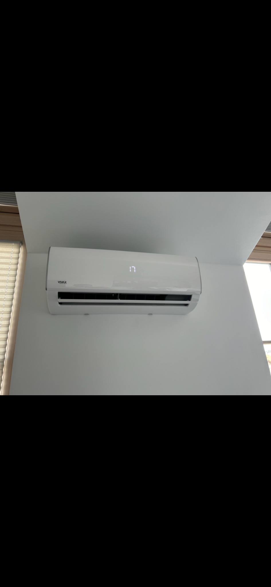 Biały klimatyzator marki Vivax zamontowany na białej ścianie, wyświetlający temperaturę 17 stopni, z widocznymi żaluzjami okiennymi w tle.