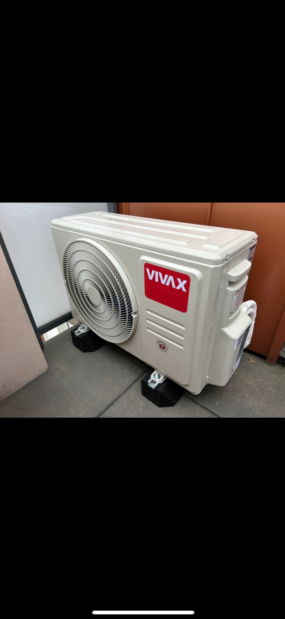 Zewnętrzna jednostka klimatyzacji VIVAX, ustawiona na czarnych, gumowych podkładkach antywibracyjnych na szarym betonowym podłożu, widoczna ściana budynku w tle.