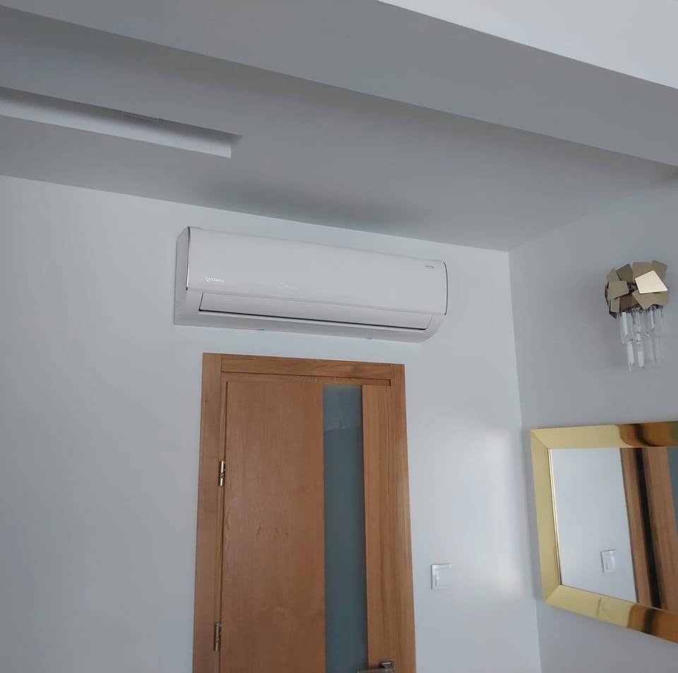 Biała jednostka klimatyzacji zamontowana nad drewnianymi drzwiami z szybą w nowoczesnym wnętrzu z dekoracyjnym oświetleniem i złotym lustrem.