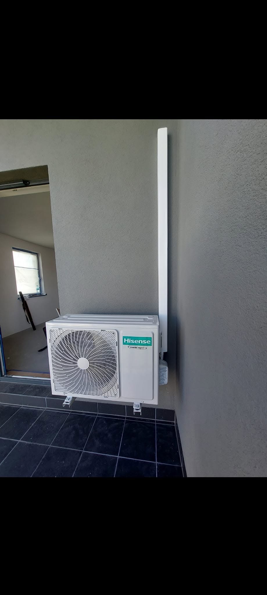 Biała jednostka zewnętrzna klimatyzatora Hisense zamontowana na szarej ścianie, widoczne czarne płytki podłogowe i fragment wnętrza z oknem.