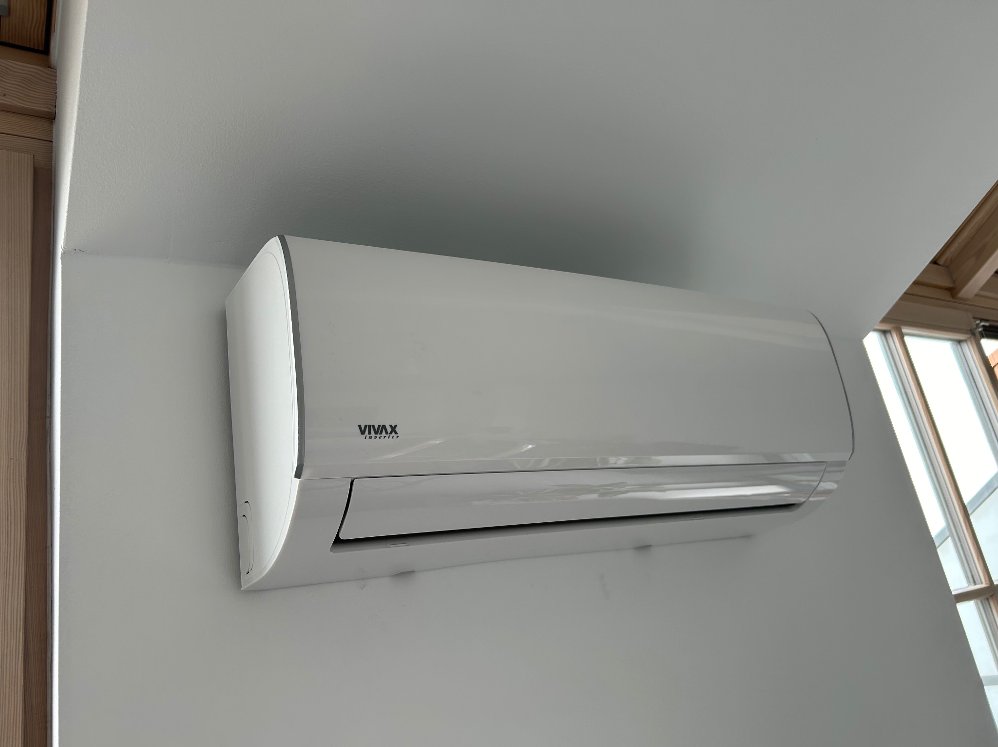Biała klimatyzacja VIVAX Inverter zamontowana na ścianie w jasnym pomieszczeniu, widoczne okno z drewnianą ramą.
