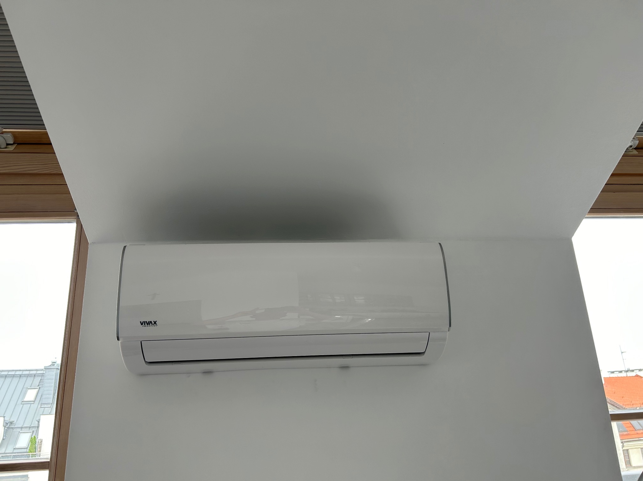 Biała jednostka wewnętrzna klimatyzacji marki Vivax, zamontowana na białej ścianie w pomieszczeniu z oknem dachowym. Widok na budynki na zewnątrz.