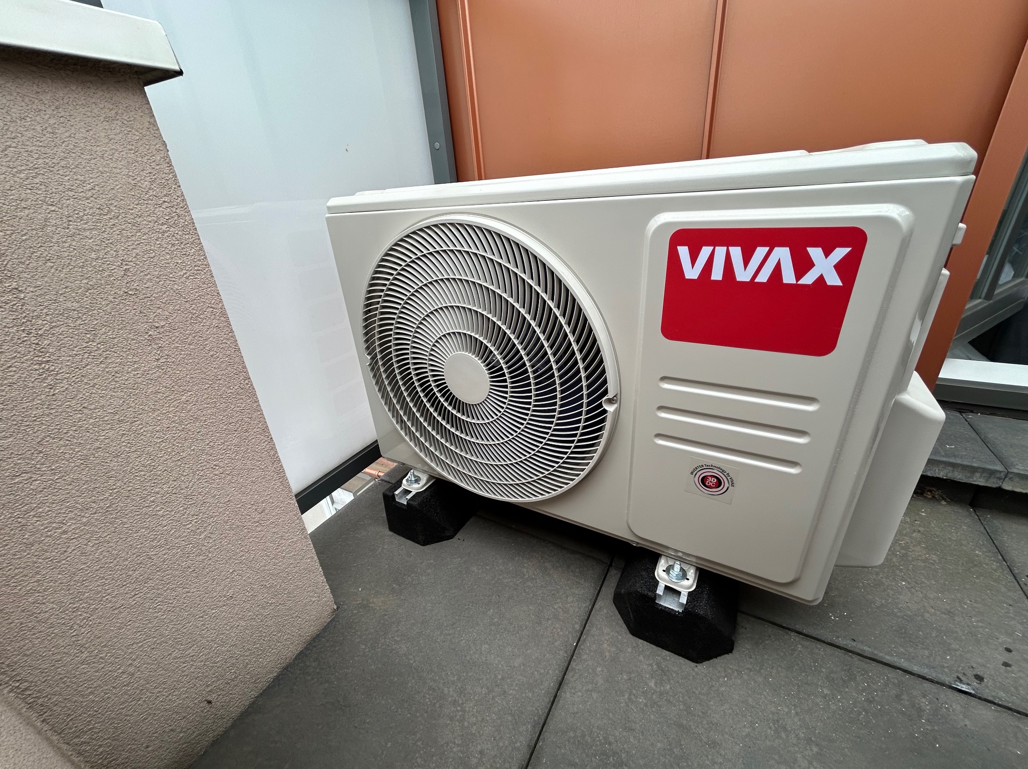 Zewnętrzna jednostka klimatyzatora marki Vivax, zamontowana na czarnych podkładkach antywibracyjnych na szarym, betonowym podłożu, z widoczną ścianą budynku w tle.