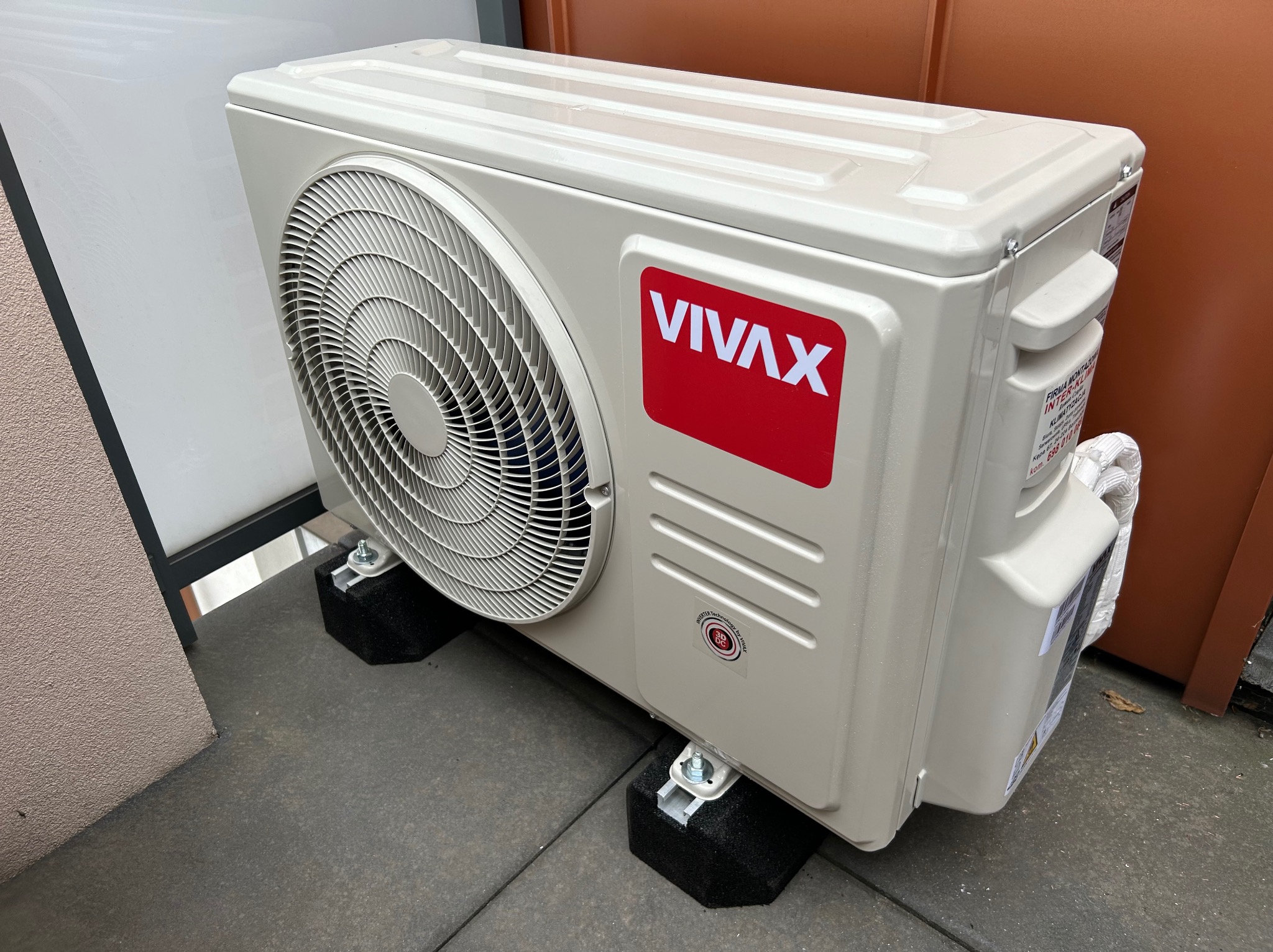 Zewnętrzna jednostka klimatyzatora VIVAX umieszczona na czarnych, tłumiących drgania podkładkach z metalowymi wspornikami, na szarym betonowym podłożu, z widocznymi elementami elewacji budynku w tle.