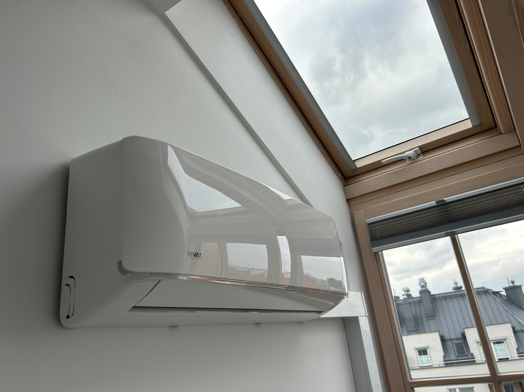 Nowoczesny, biały klimatyzator marki Vivax zamontowany na skośnej ścianie pod oknem dachowym. Widok na budynki przez okno.