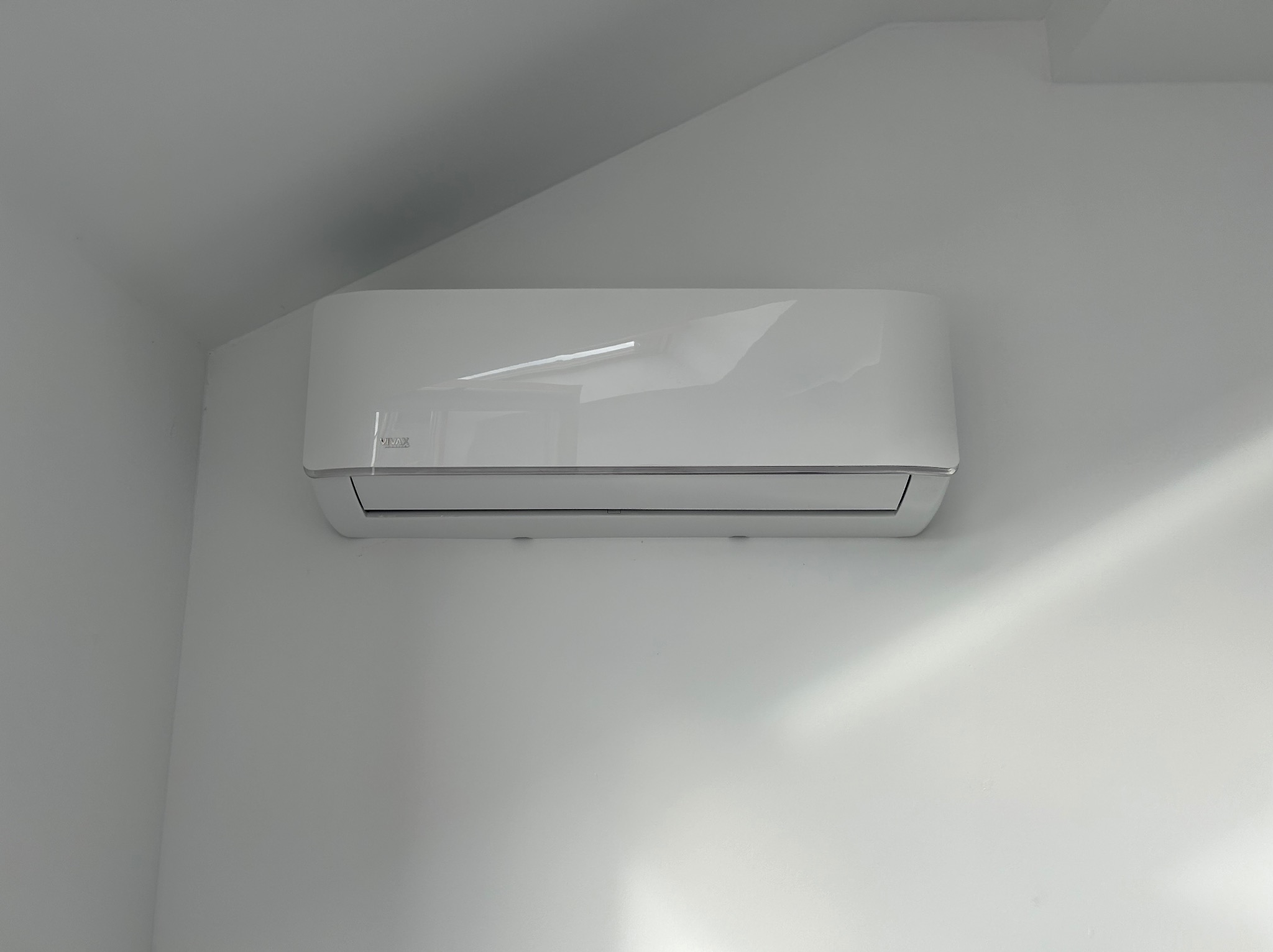 Biała jednostka wewnętrzna klimatyzatora marki Vivax zamontowana na białej ścianie pod skosem dachu.