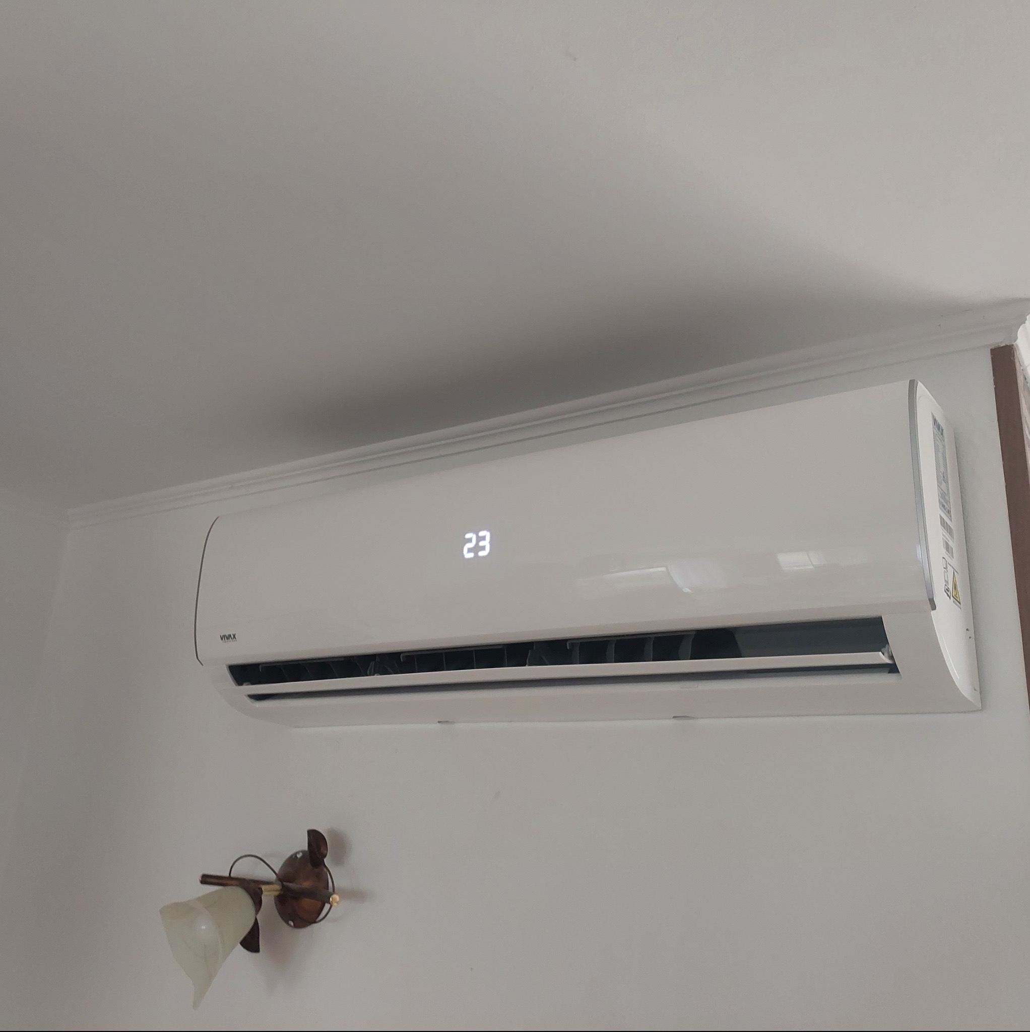 Biała jednostka klimatyzacji VIVAX zamontowana na ścianie w pomieszczeniu, wyświetlająca temperaturę 23 stopnie, widoczna lampka ścienna poniżej.