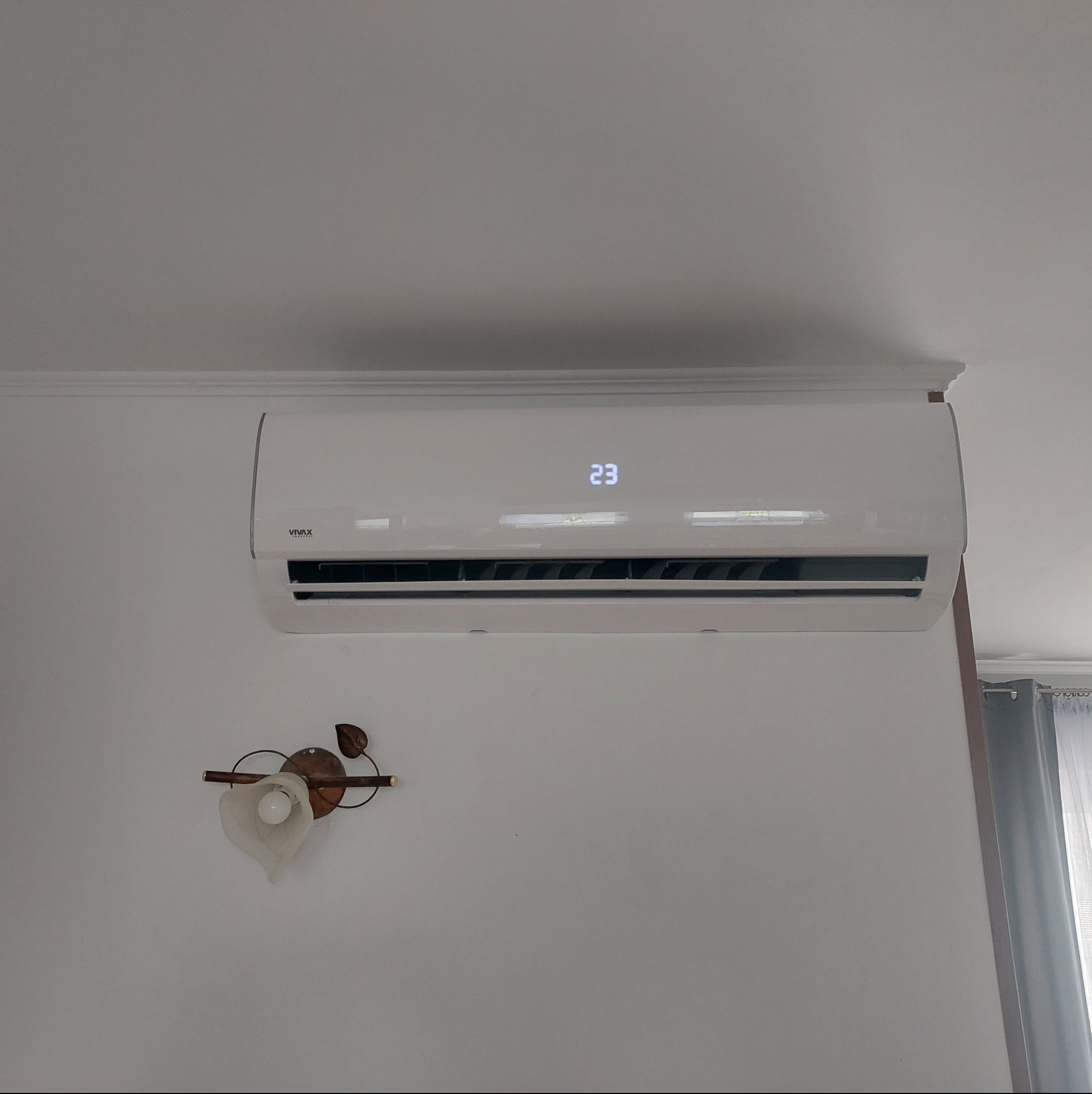 Biała klimatyzacja marki Vivax zamontowana na ścianie w pomieszczeniu, wyświetlająca temperaturę 23 stopnie Celsjusza, obok lampka w stylu retro.