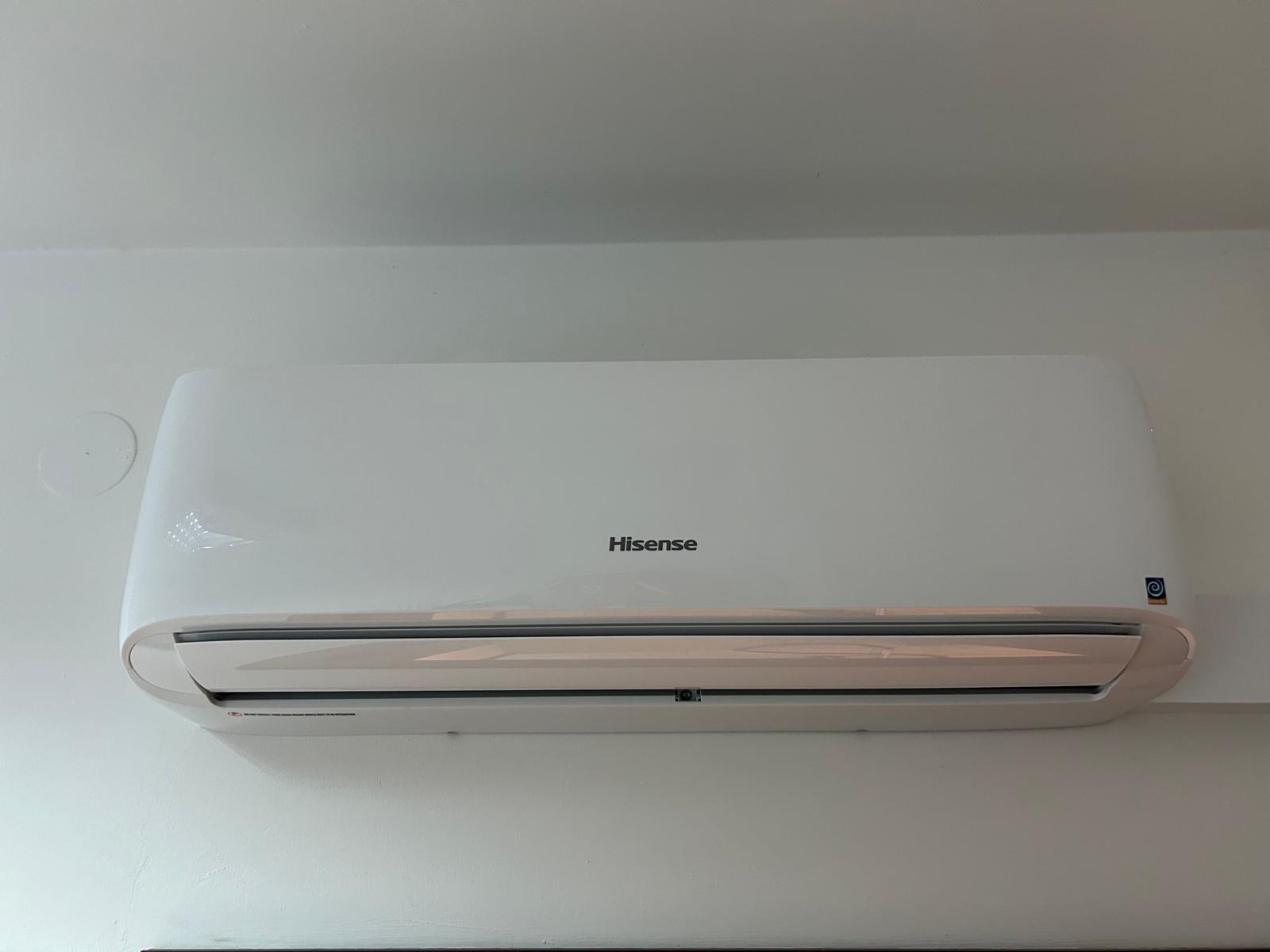 Biały klimatyzator Hisense zamontowany na białej ścianie, widoczny z dołu, z delikatnym światłem odbijającym się od obudowy.