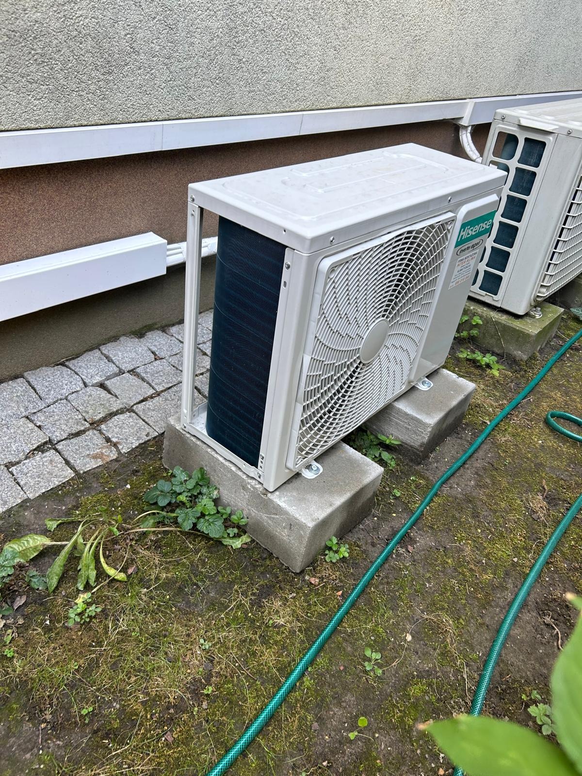 Dwa białe, zewnętrzne moduły klimatyzacji Hisense, ustawione na betonowych blokach przy ścianie budynku z brązową elewacją, widoczne zarośla i zielony wąż ogrodowy.