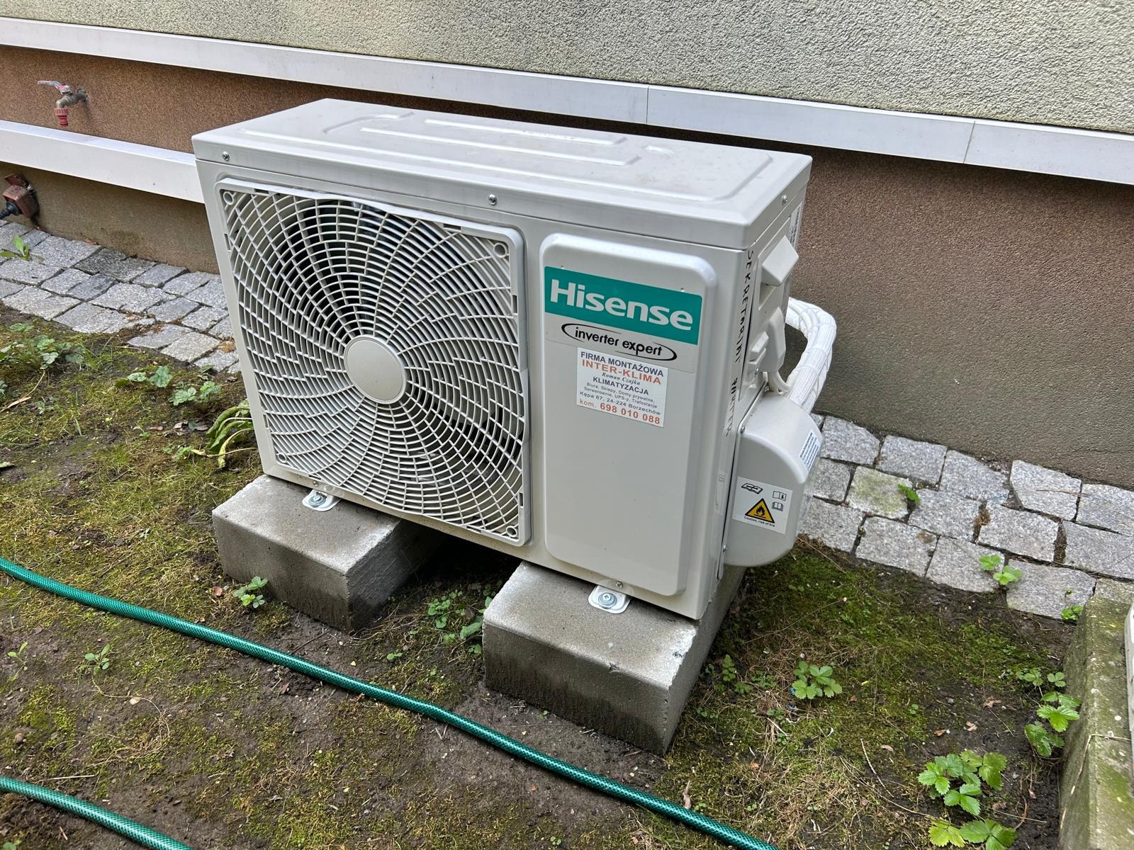 Zewnętrzna jednostka klimatyzacji Hisense zamontowana na dwóch betonowych bloczkach na trawniku, z widoczną naklejką firmy montażowej Inter-Klima i zielonym wężem ogrodowym w tle.