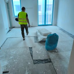 Adrian Grobelski - Pracownik w kamizelce odblaskowej przechodzi przez pomieszczenie z przygotowaną podłogą, zabezpieczone okna folią, widoczny worek z odpadami.