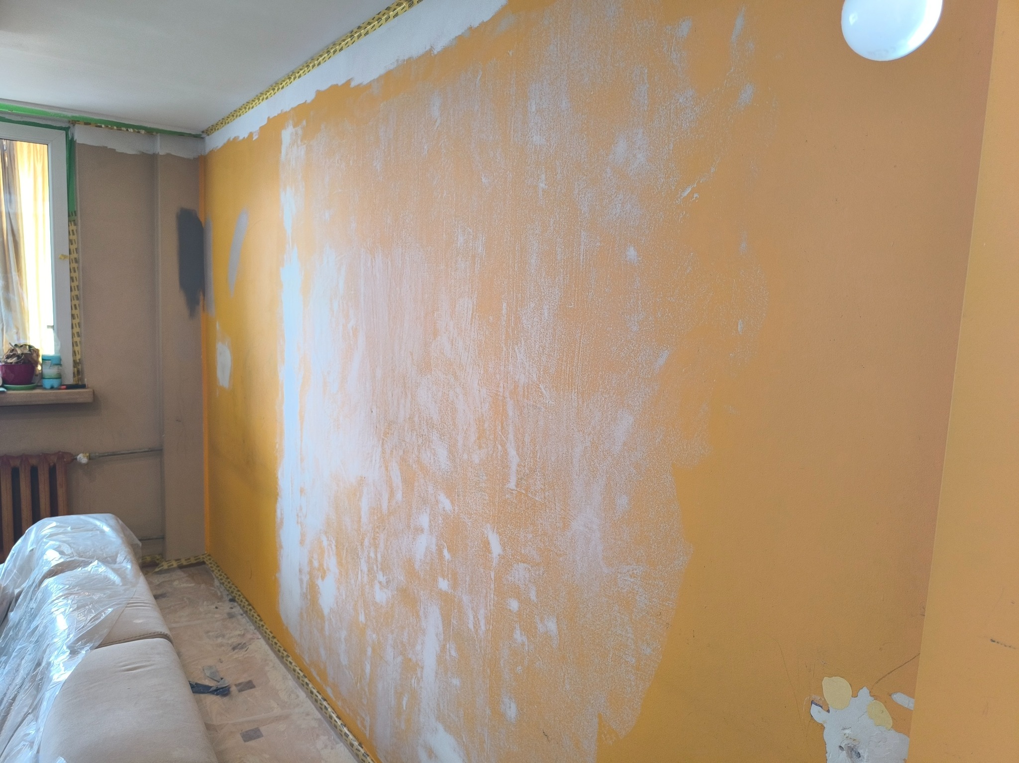 Pomarańczowa ściana w trakcie szpachlowania, widoczne ślady białej gładzi, zabezpieczona taśmą malarską pod sufitem i przy podłodze, meble przykryte folią ochronną.