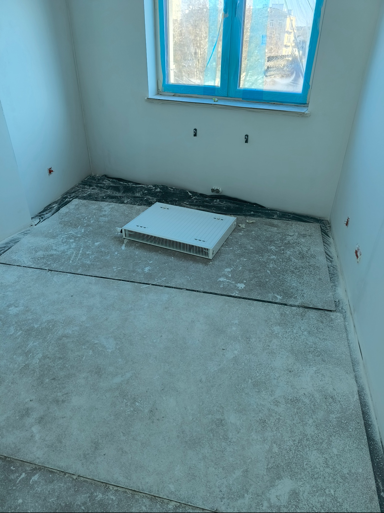 Pokój w trakcie prac wykończeniowych: podłoga pokryta płytami, grzejnik na podłodze, okno zabezpieczone folią, ściany pomalowane na biało, widoczne ślady szpachli.