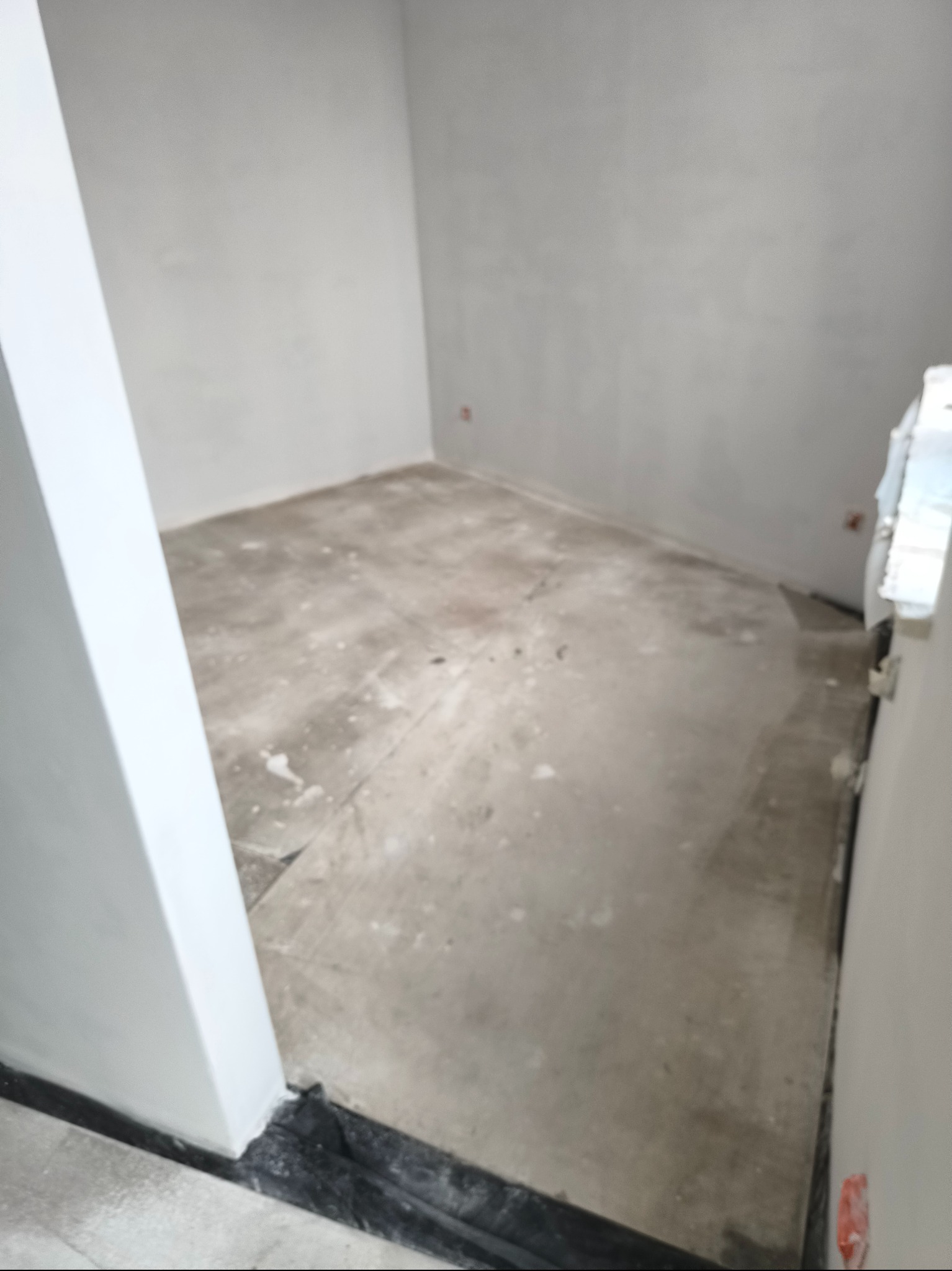 Pokój w stanie surowym: ściany po szpachlowaniu, podłoga przygotowana do dalszych prac wykończeniowych, widoczne zabezpieczenia folią przy ścianach.