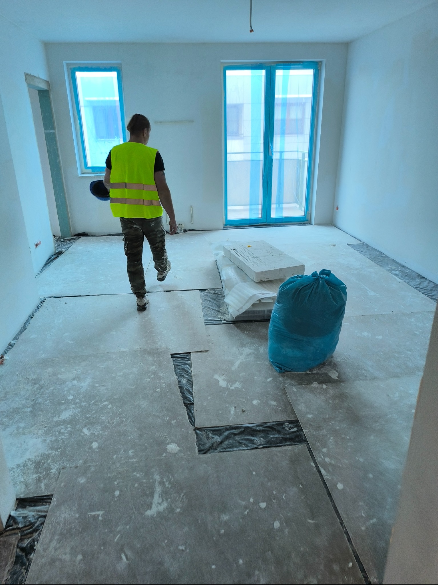 Pracownik w kamizelce odblaskowej przechodzi przez pomieszczenie z przygotowaną podłogą, zabezpieczone okna folią, widoczny worek z odpadami.
