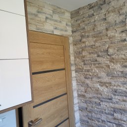 Rafal Nowicki - Ściana wykończona dekoracyjnym kamieniem w odcieniach beżu i szarości, sąsiadująca z drewnianymi drzwiami z ciemnymi akcentami i fragmentem białej szafki kuchennej.