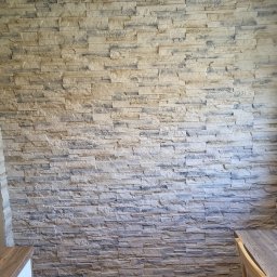 Rafal Nowicki - Ściana wyłożona dekoracyjnym kamieniem w odcieniach beżu i szarości, ułożonym warstwowo, widoczny fragment drewnianej podłogi i narożnik białej szafki oraz krzesło.