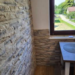 Rafal Nowicki - Fragment wnętrza z kamienną okładziną ściany w odcieniach beżu i szarości, widok na okno z widokiem na zielony ogród i dom z czerwonym dachem, fragment blatu stołu z podkładką.