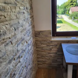 Fragment wnętrza z kamienną okładziną ściany w odcieniach beżu i szarości, widok na okno z widokiem na zielony ogród i dom z czerwonym dachem, fragment blatu stołu z podkładką.