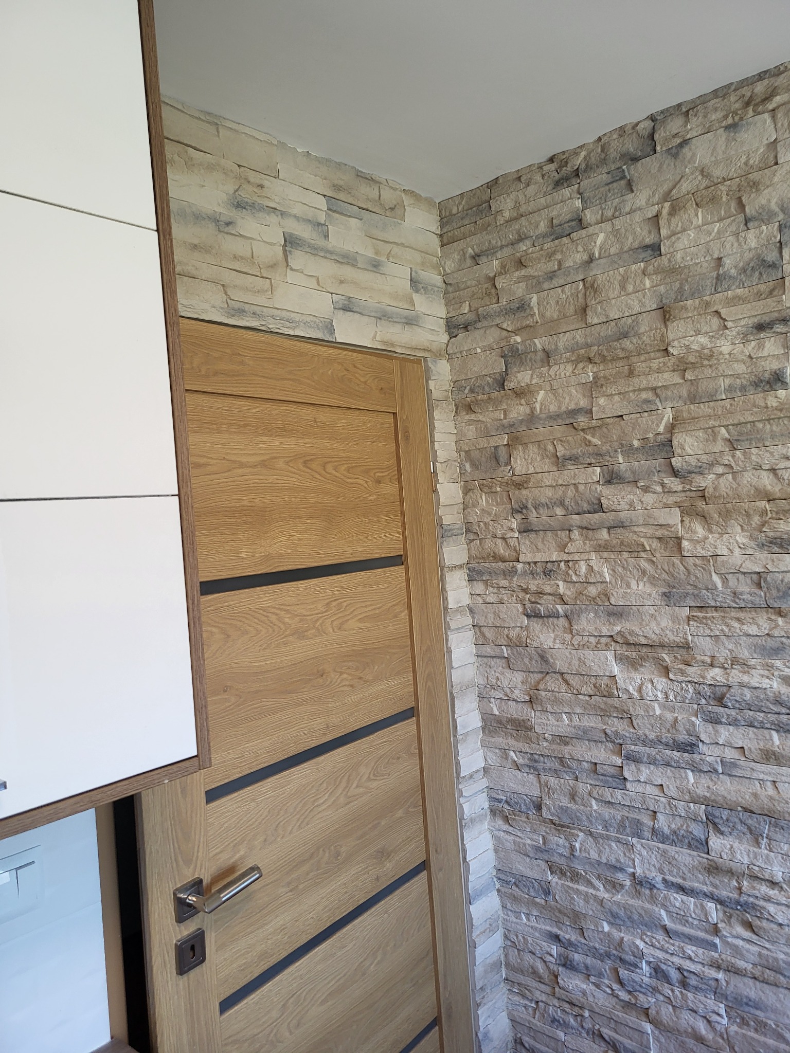 Ściana wykończona dekoracyjnym kamieniem w odcieniach beżu i szarości, sąsiadująca z drewnianymi drzwiami z ciemnymi akcentami i fragmentem białej szafki kuchennej.