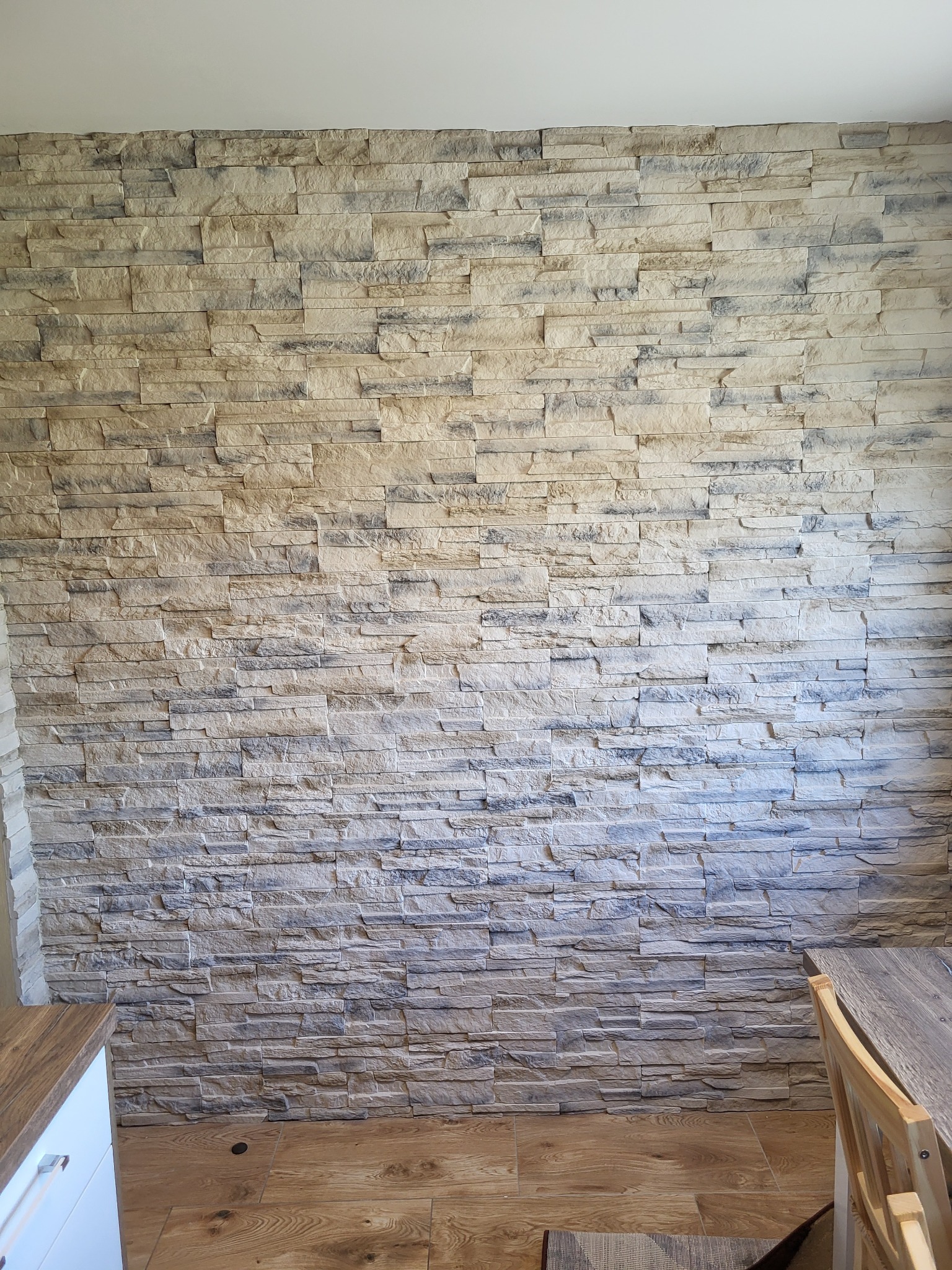 Ściana wyłożona dekoracyjnym kamieniem w odcieniach beżu i szarości, ułożonym warstwowo, widoczny fragment drewnianej podłogi i narożnik białej szafki oraz krzesło.