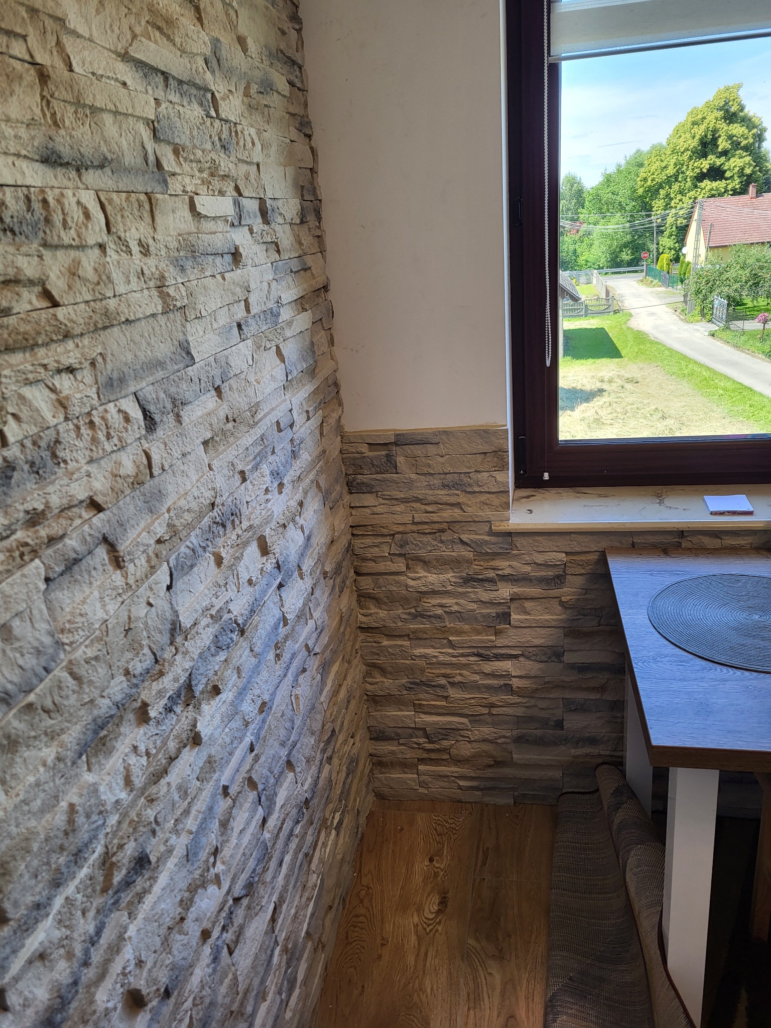 Fragment wnętrza z kamienną okładziną ściany w odcieniach beżu i szarości, widok na okno z widokiem na zielony ogród i dom z czerwonym dachem, fragment blatu stołu z podkładką.