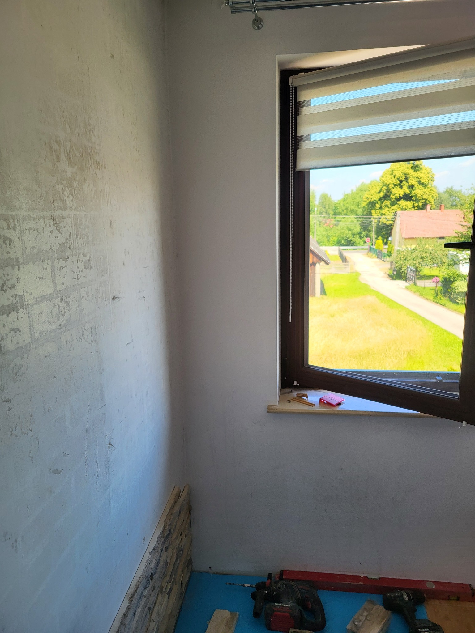 Ściana w trakcie wykańczania z częściowo położoną okładziną kamienną, narzędzia glazurnicze na podłodze zabezpieczonej folią, widok z okna na zewnątrz.