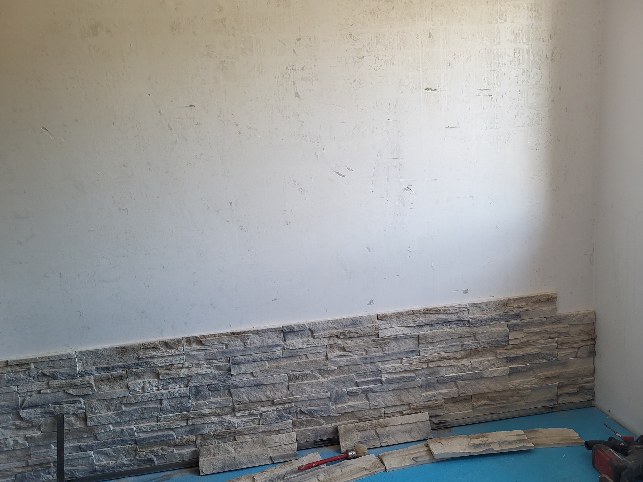 Montaż dekoracyjnej okładziny kamiennej na ścianie. Częściowo wykończona ściana z szarej okładziny, na podłodze leżą płytki i czerwony pędzel, widoczny także fragment niezagruntowanej ściany.