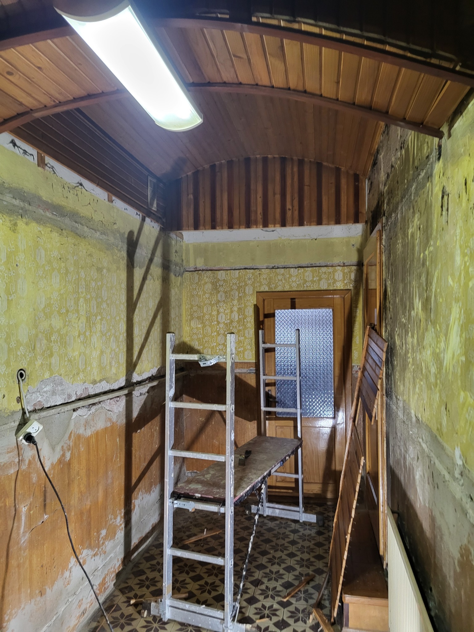 Wnętrze w trakcie remontu: odsłonięte fragmenty ścian z resztkami tapety, drewniany sufit, rozstawiony aluminiowy ruszt, widoczne kable elektryczne i stara podłoga z wzorem.