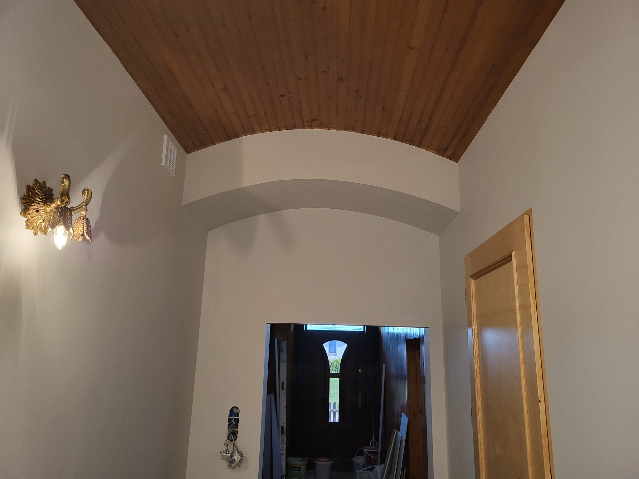 Wnętrze z drewnianym sufitem, łukowym przejściem i ozdobnym, złotym kinkietem w stylu retro, widoczne puszki z farbą i niedokończone gniazdko elektryczne.