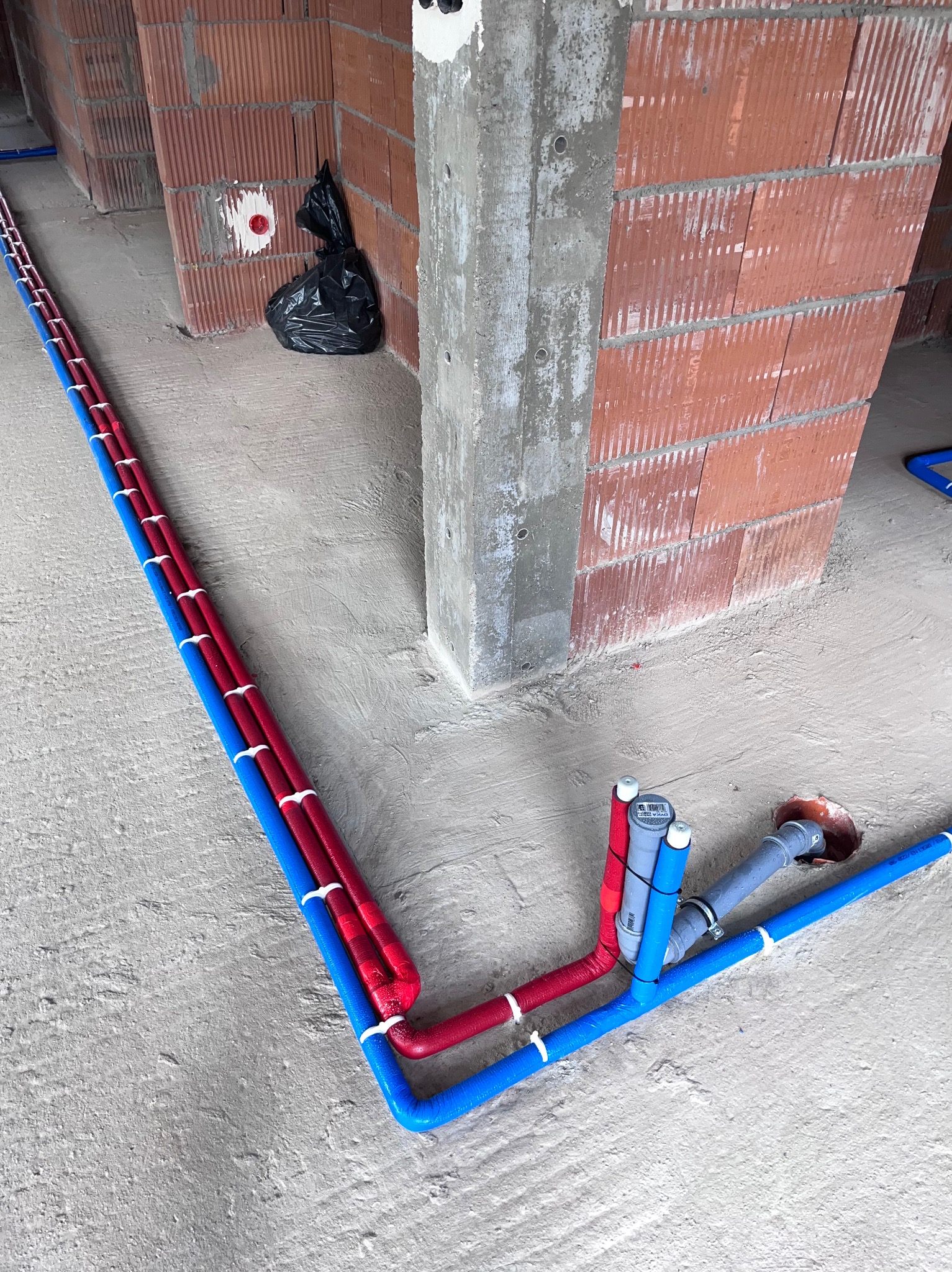 Wnętrze w stanie surowym: Ułożone równolegle rury instalacji wodnej w kolorach niebieskim i czerwonym, przymocowane do ściany klinkierowej za pomocą białych uchwytów, widoczne przyłącza i fragment...