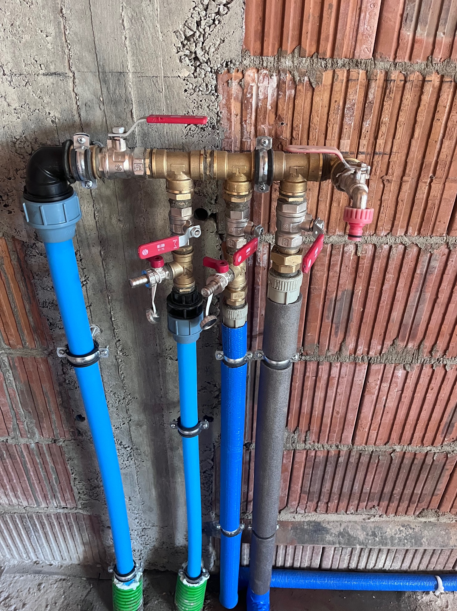 Rozdzielacz instalacji wodnej z niebieskimi rurami PEX i zaworami z czerwonymi rączkami, zamocowany na ścianie z cegły i surowego betonu.