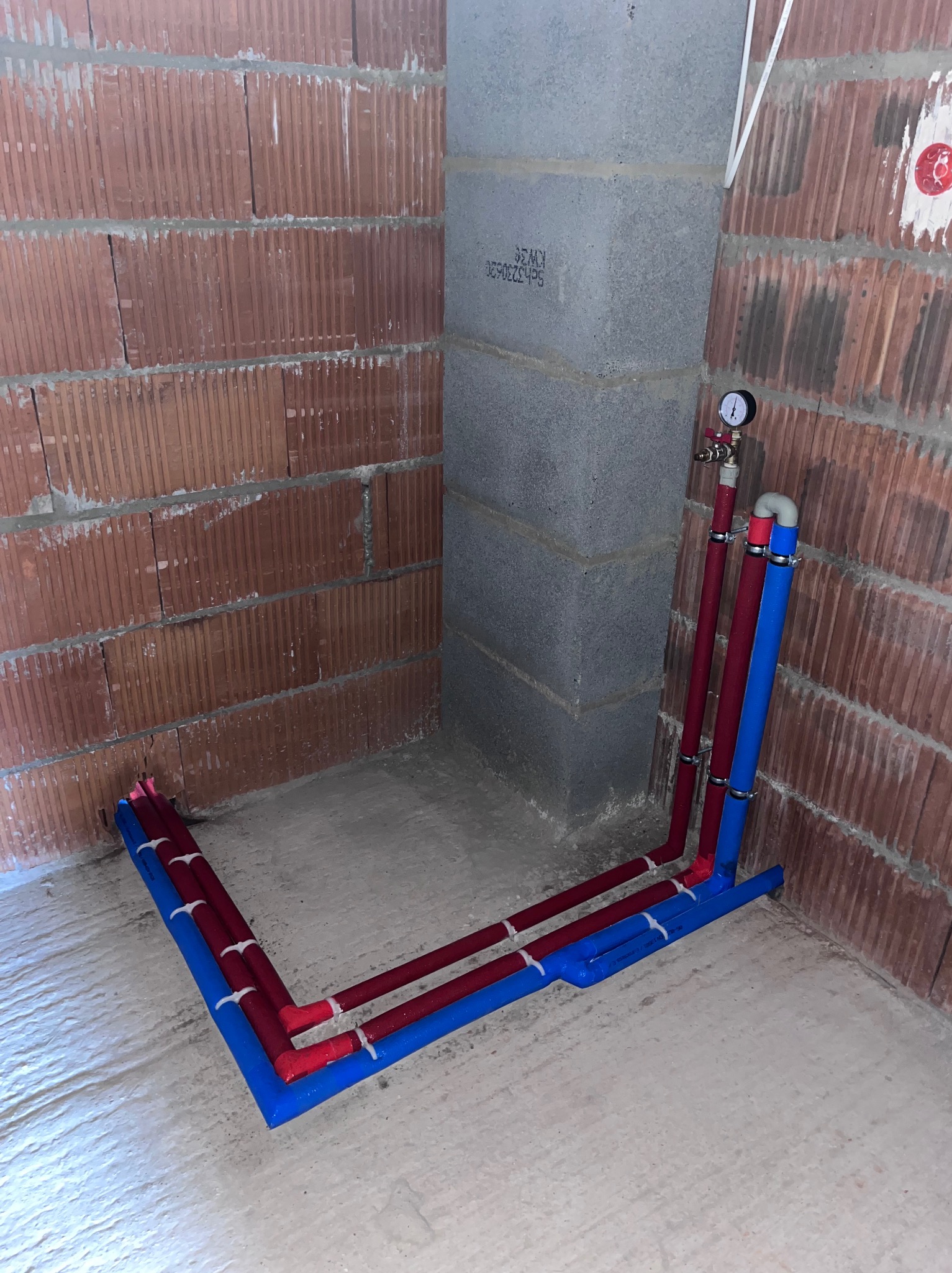 Instalacja hydrauliczna w trakcie montażu w surowym stanie, widoczne rury czerwone i niebieskie, manometr oraz ściany z cegły dziurawki i bloczków betonowych.
