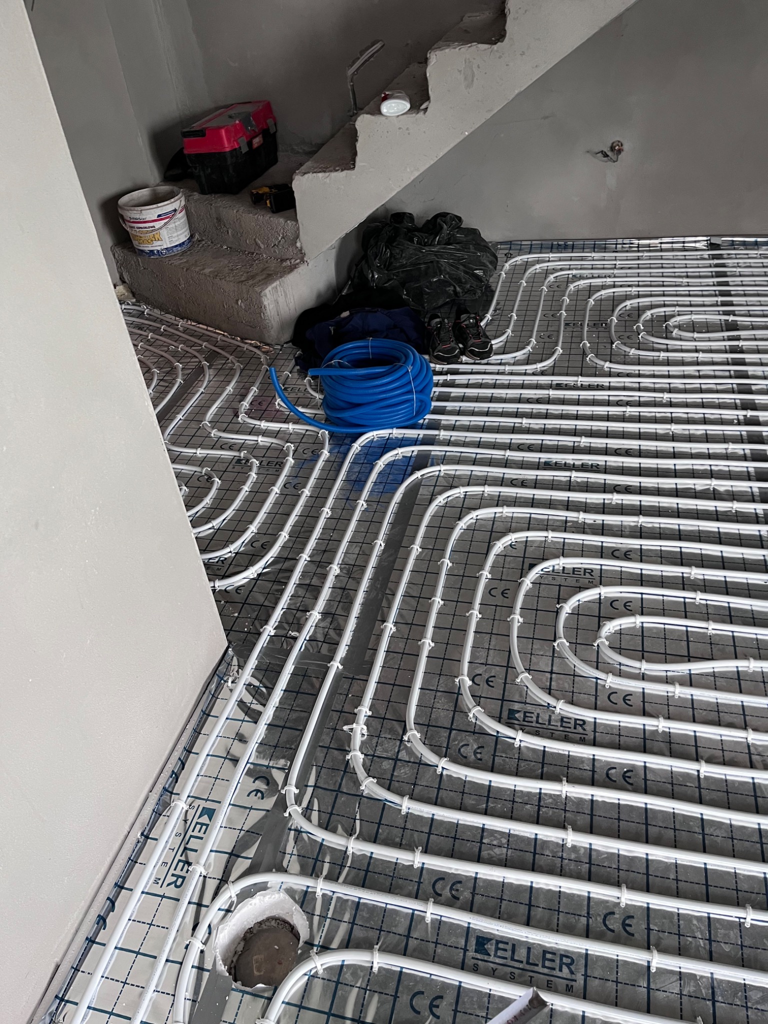 Białe rury ogrzewania podłogowego ułożone na folii z nadrukiem, widoczny fragment schodów w surowym stanie, narzędzia i zwój niebieskiej rury