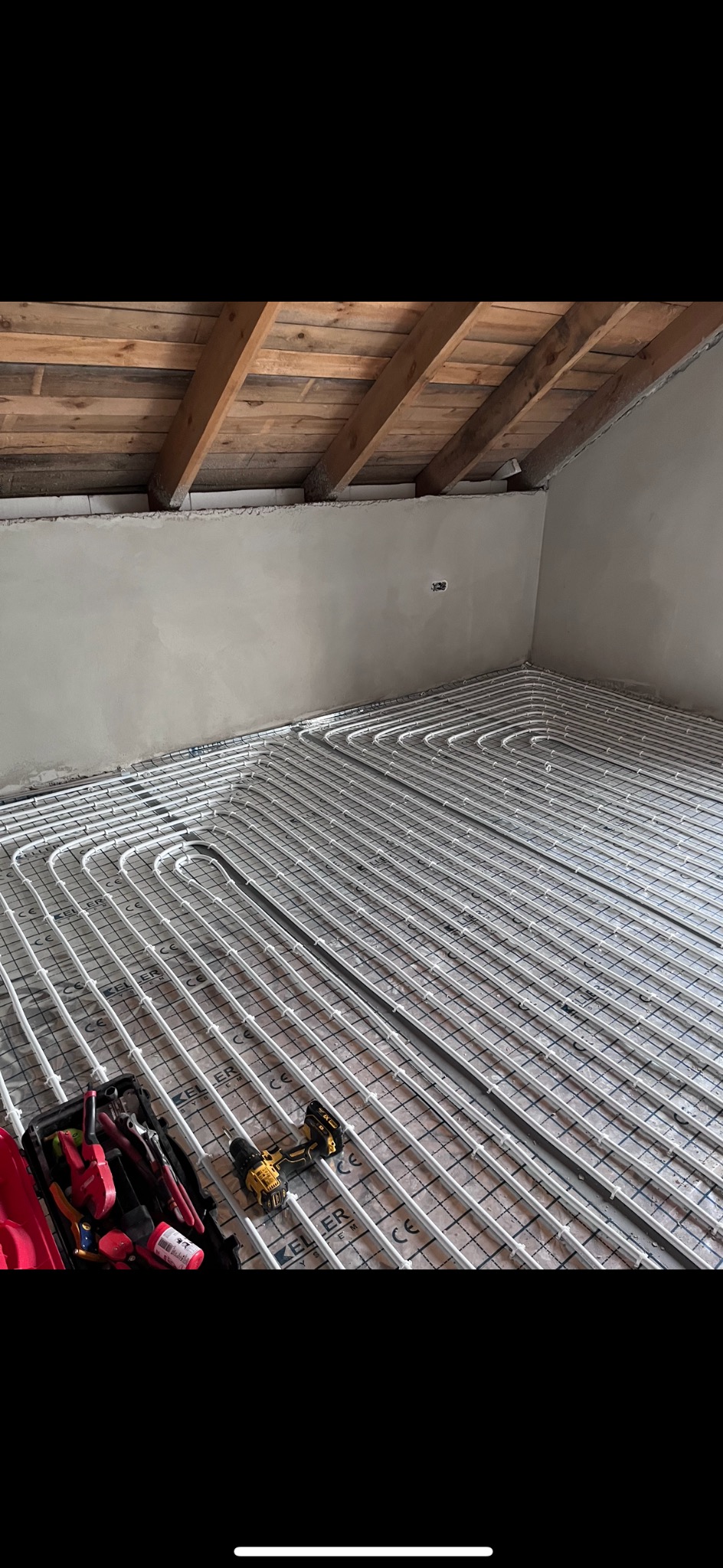 Białe rurki ogrzewania podłogowego ułożone na izolacji w surowym pomieszczeniu z drewnianym stropem, widoczna skrzynka z narzędziami i wkrętarka.