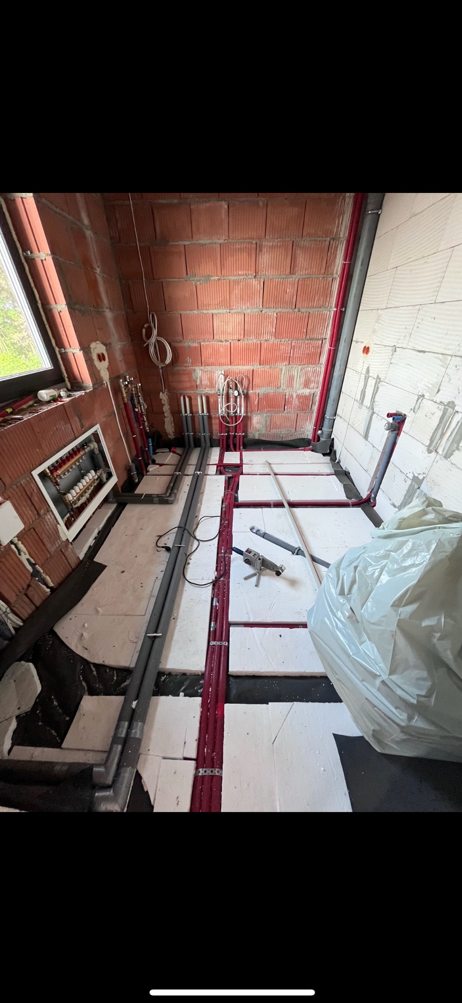 Wnętrze w trakcie montażu ogrzewania podłogowego z widocznymi czerwonymi rurami mocowanymi do izolacji, szarymi rurami przy ścianie, rozdzielaczem oraz narzędziami do zgrzewania rur.