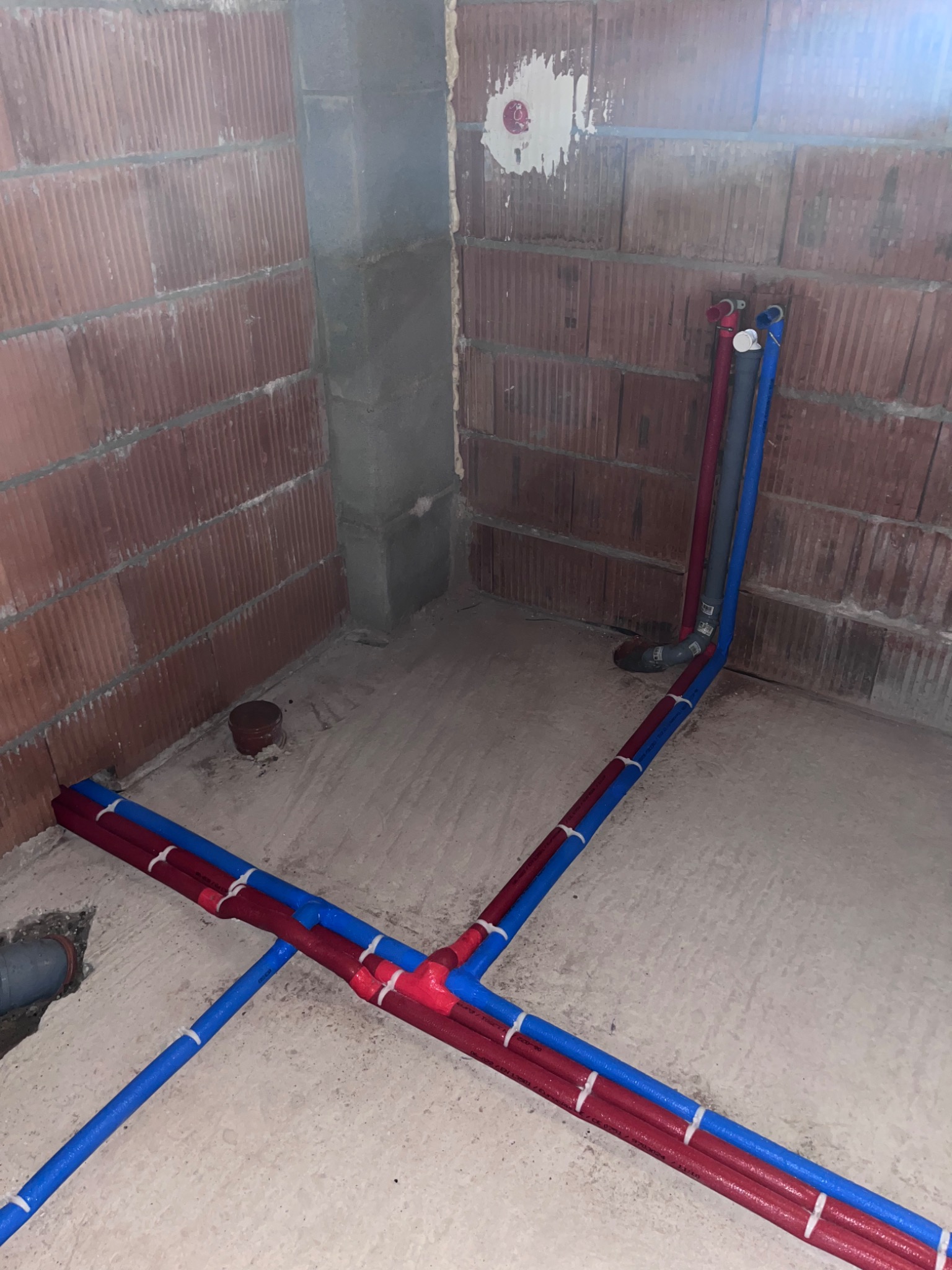 Surowe wnętrze z instalacją wodną w trakcie montażu: czerwone i niebieskie rury prowadzone po podłodze i w górę ściany z cegły, połączone białymi opaskami zaciskowymi, widoczne fragmenty wylewki...