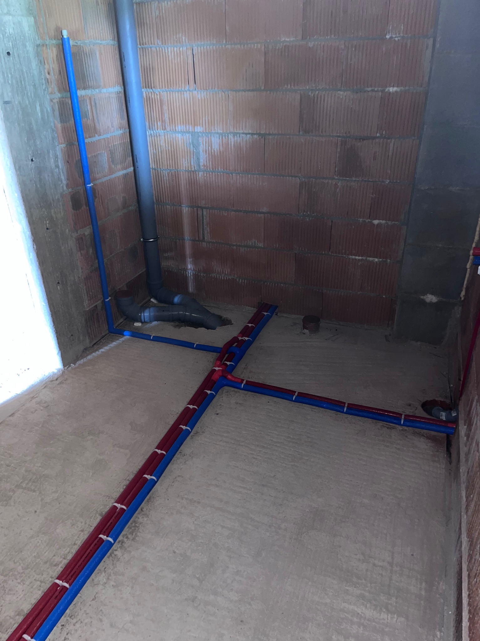 Surowy stan łazienki w trakcie prac hydraulicznych: widoczne rury kanalizacyjne i instalacyjne w kolorach niebieskim i czerwonym, przymocowane do surowej podłogi i ścian z cegły.