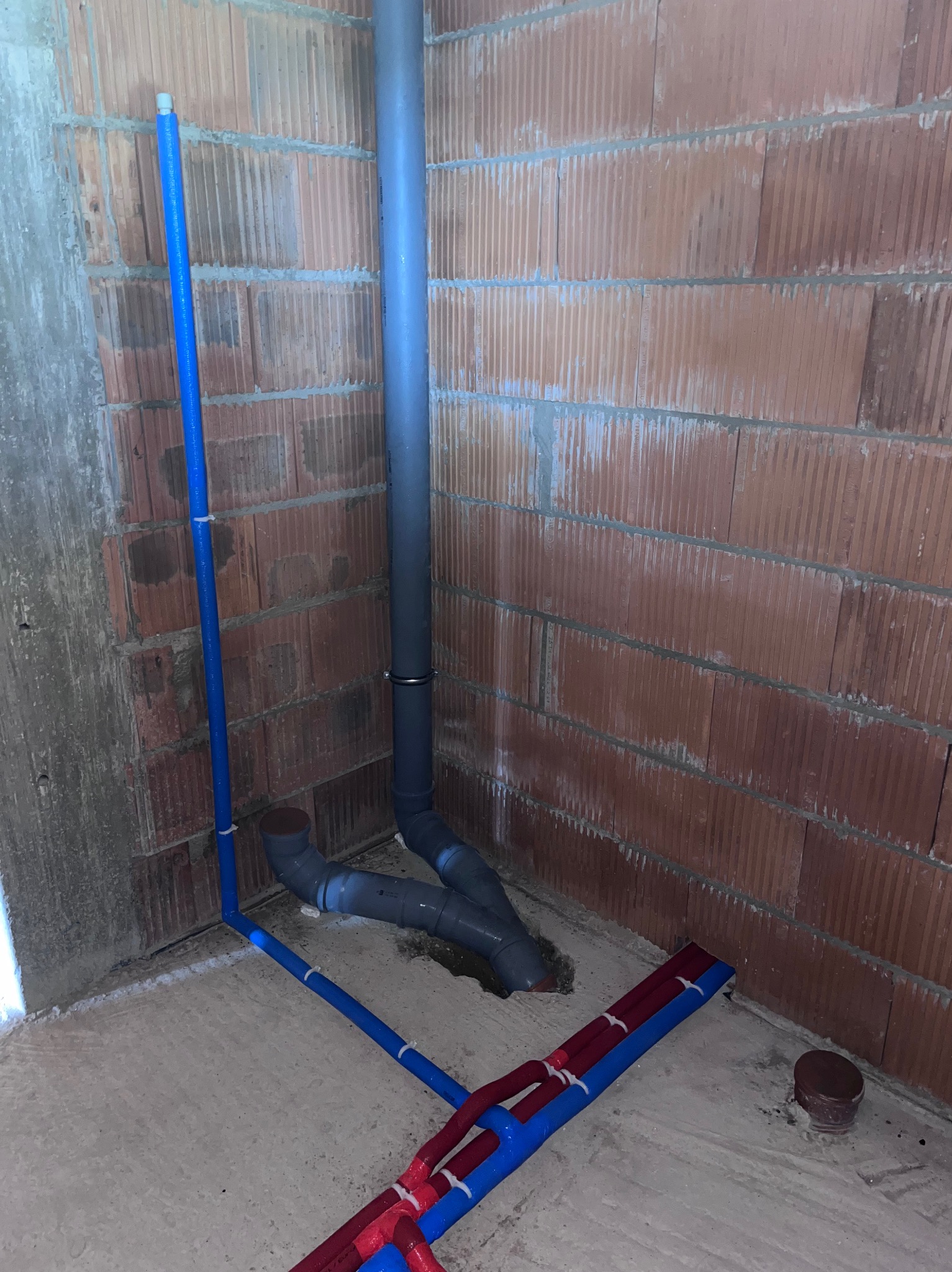 Surowe wnętrze z widocznymi elementami instalacji hydraulicznej: pionowa rura kanalizacyjna z szarego PCV, niebieskie rury wodne z tworzywa sztucznego oraz fragment ogrzewania podłogowego w kolorze...